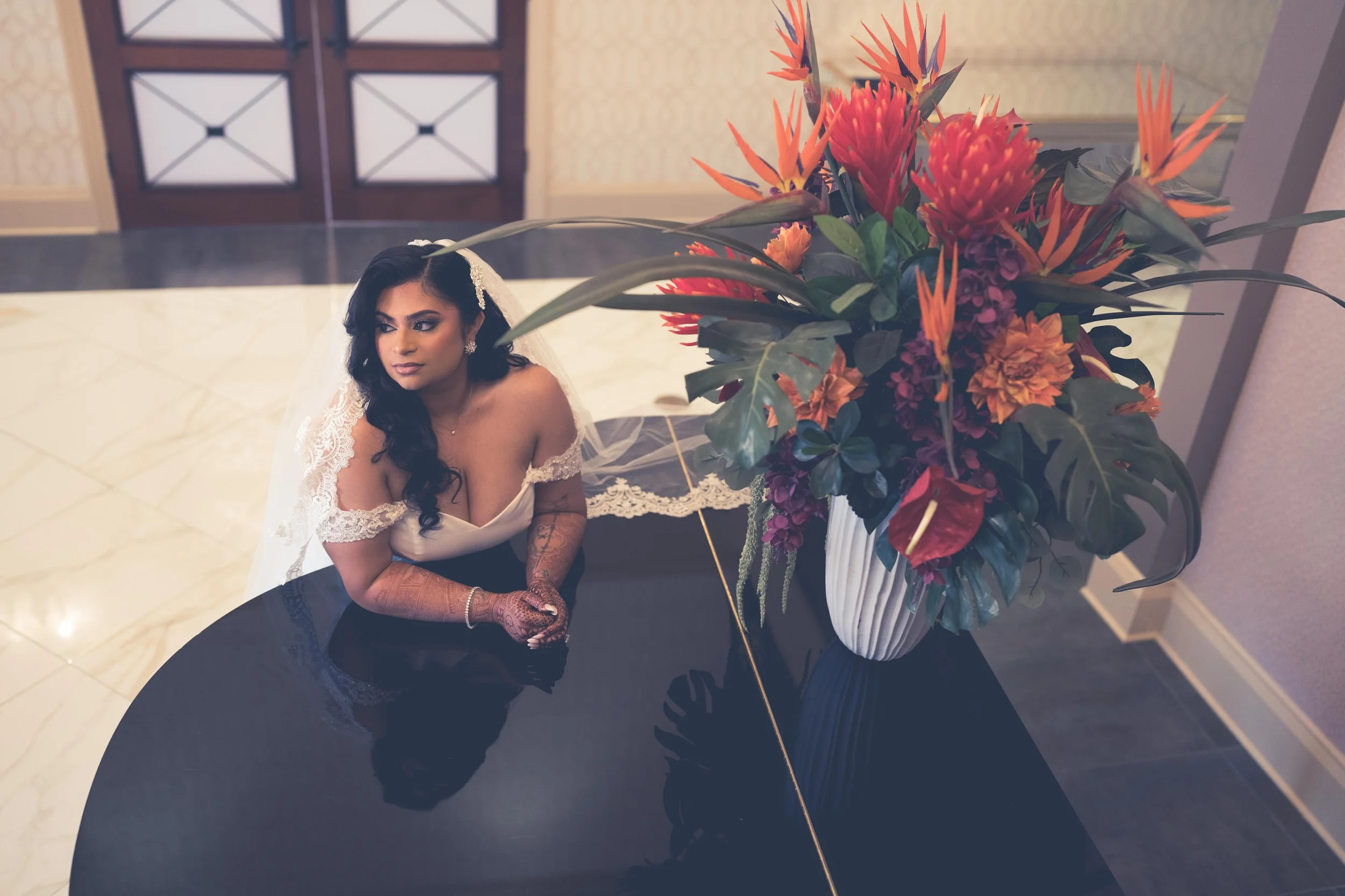 A bride with dark hair styled in loose curls, wearing a wedding dress with lace sleeves, and white gloves, sitting at a black reflective table next to a large vase of tropical flowers, in a hotel lobby.