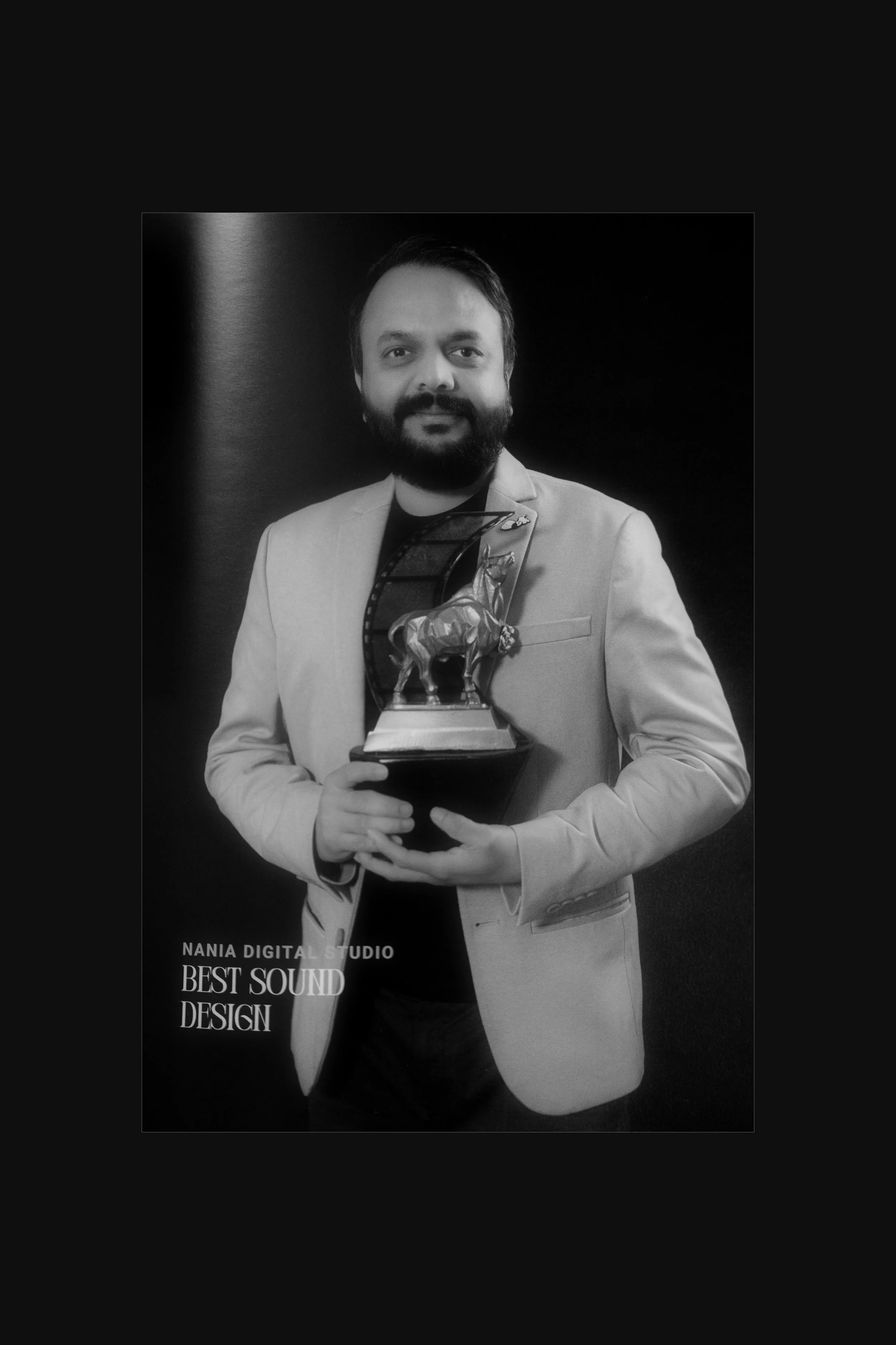 Man in a light-colored blazer holding a trophy, standing against a dark background, text on bottom left reads "Nania Digital Studio Best Sound Design."