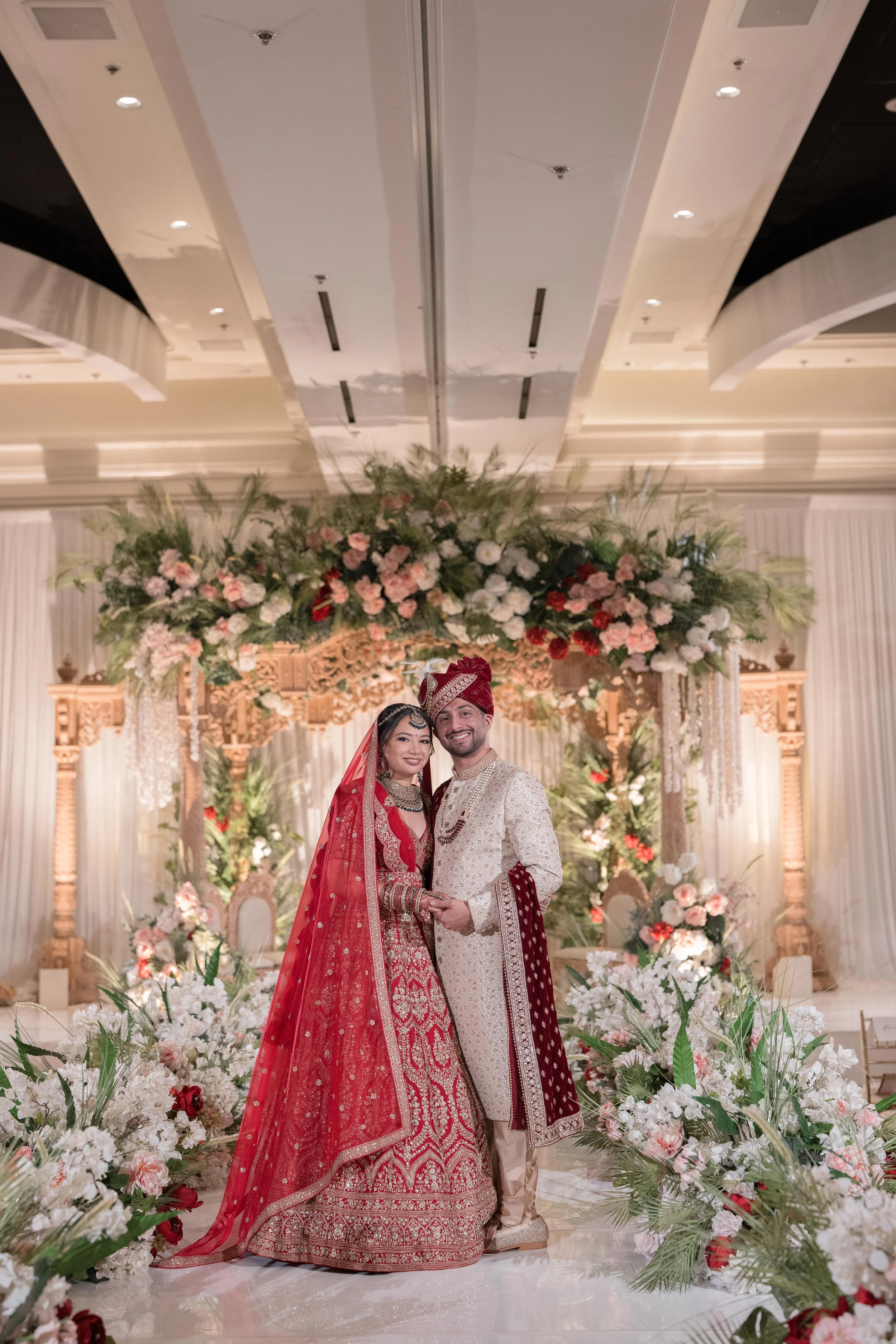 A newlywed couple in traditional Indian wedding attire standing together in front of a decorated floral arch at their wedding ceremony. The bride wears a red and gold bridal lehenga with a veil, and the groom wears a cream-colored sherwani with a red