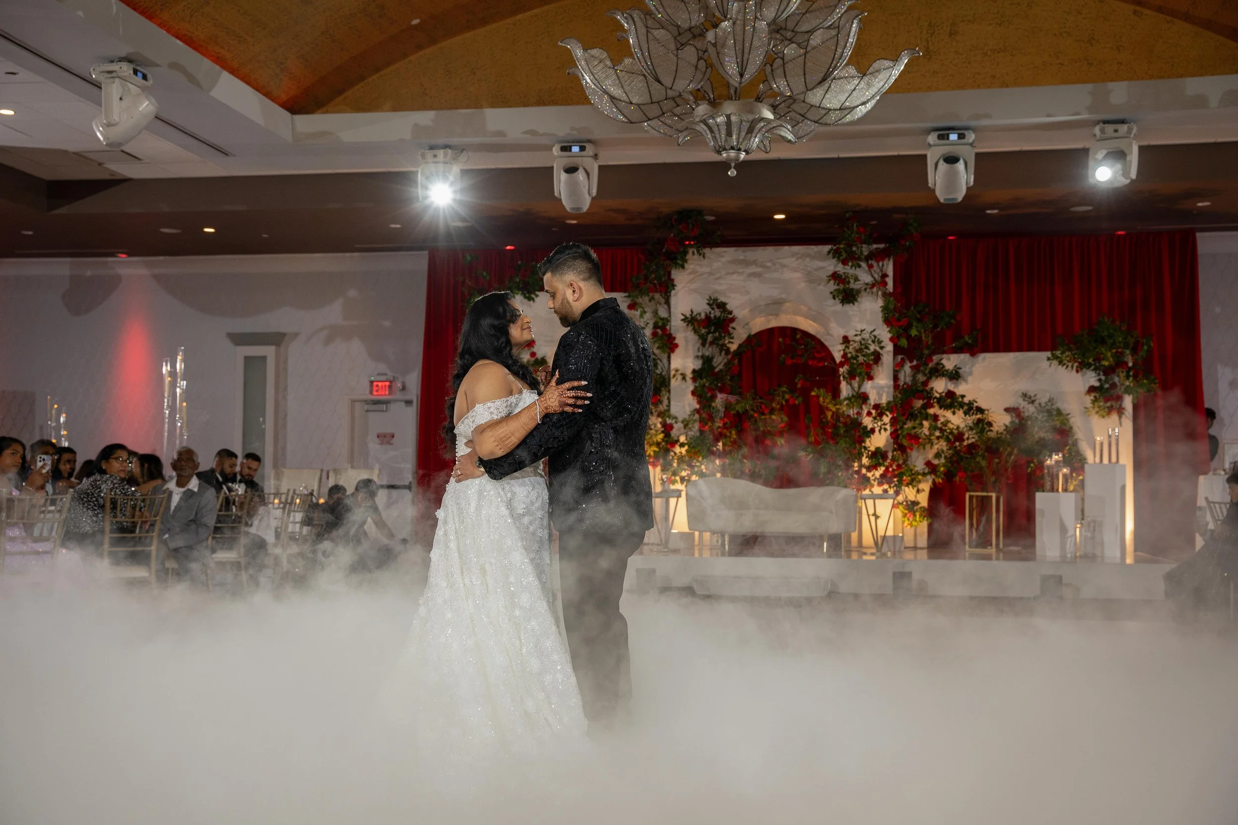 A bride and groom dancing at their wedding reception, surrounded by guests, with a decorated stage and floral backdrop in the background, and fog on the dance floor.