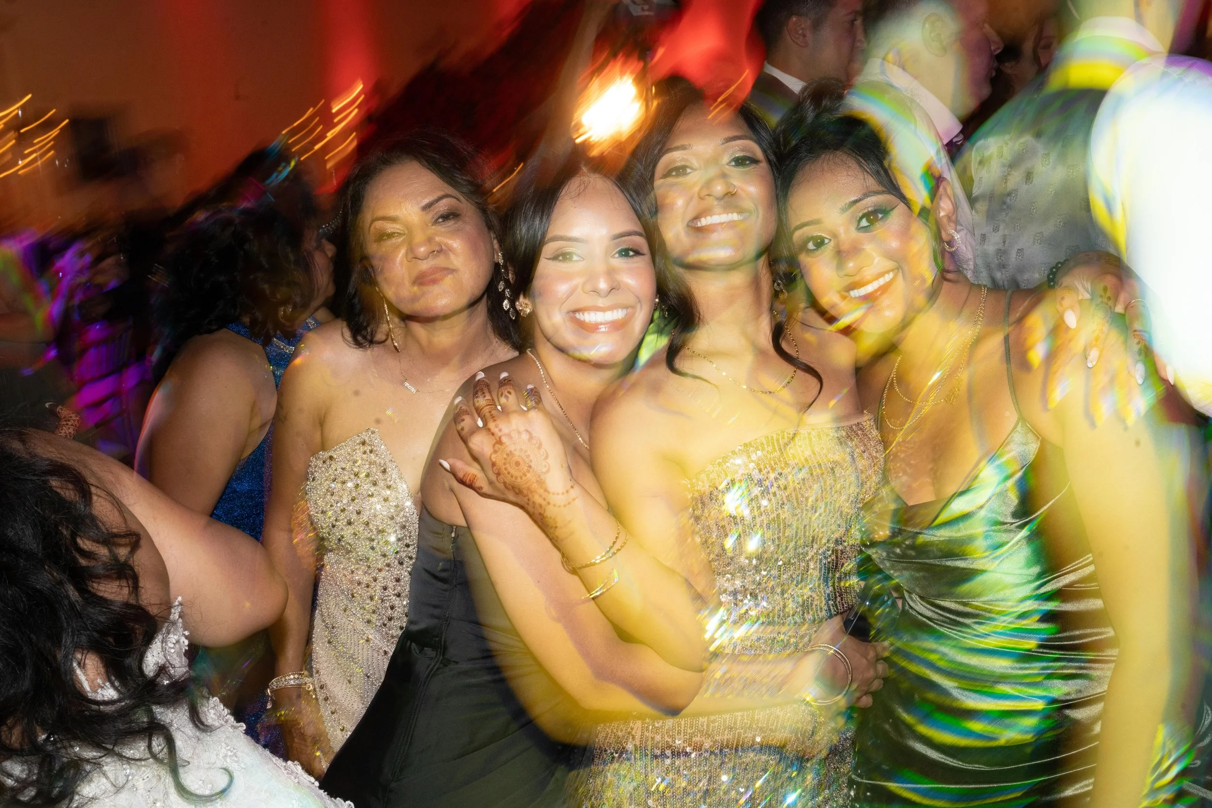 Group of women at a celebration, smiling and posing together, dressed in elegant attire with one woman wearing traditional henna on her hand.