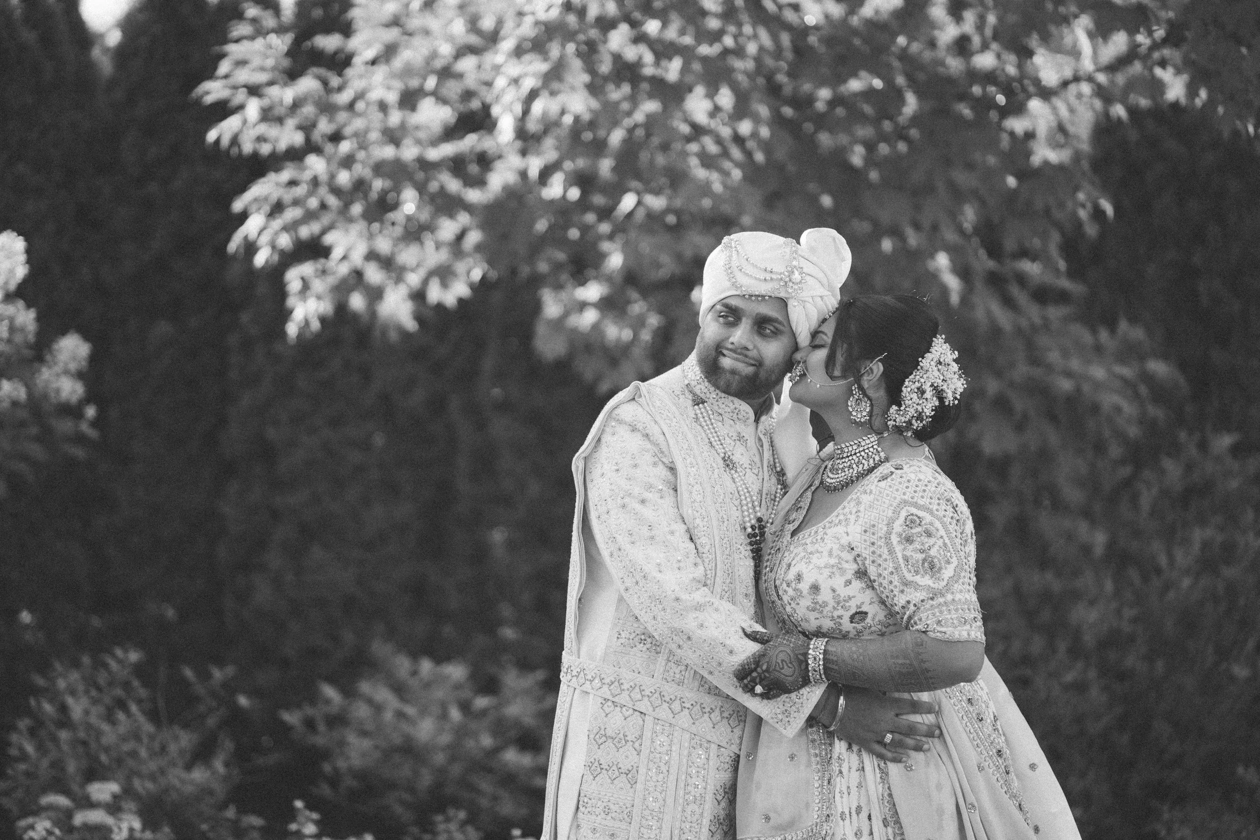 A bride and groom in traditional Indian wedding attire standing outdoors, embracing each other and smiling.