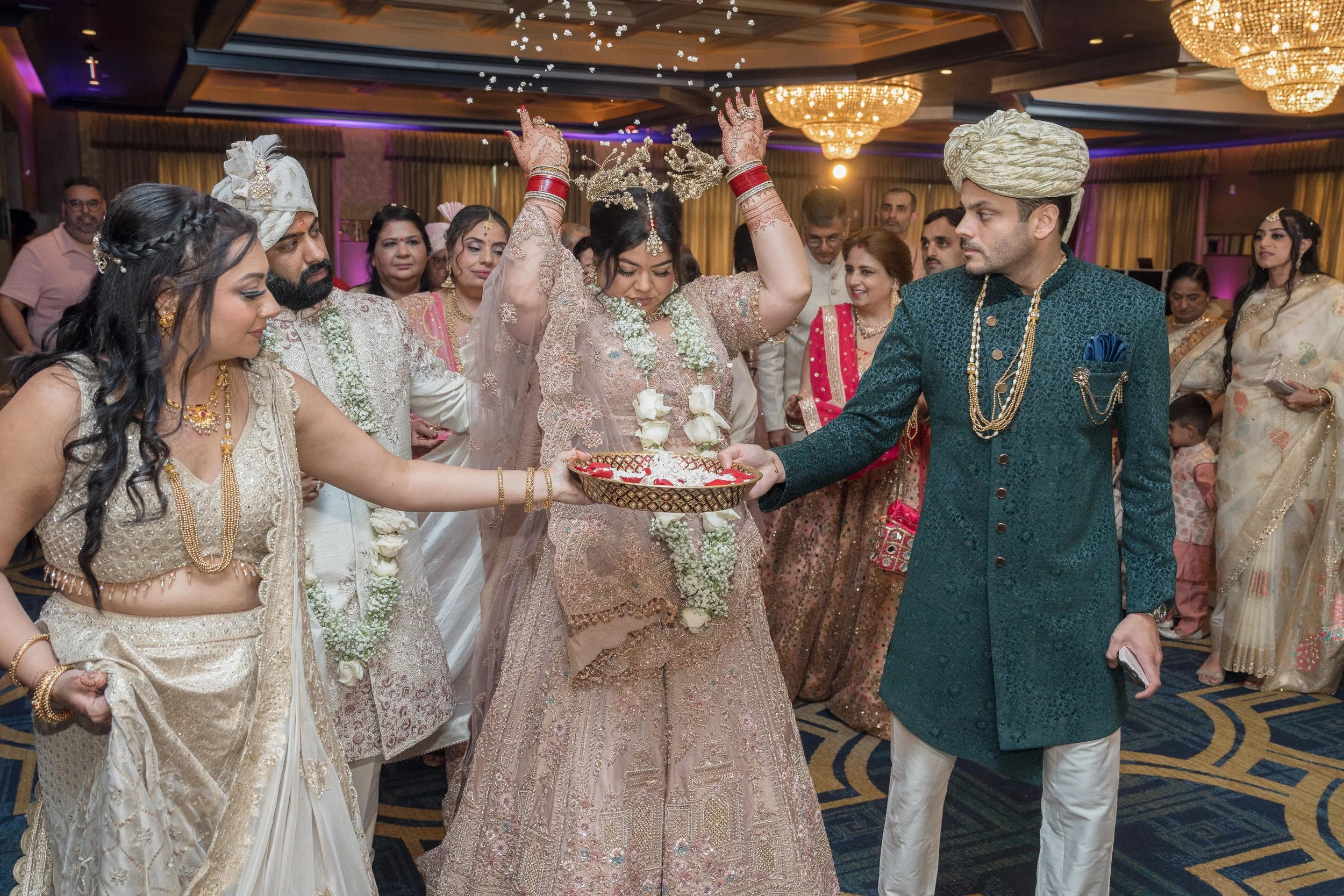 Indian wedding celebration with bride and groom in traditional attire, surrounded by family and friends, performing a ritual with flowers and a decorated tray in an ornate indoor setting.