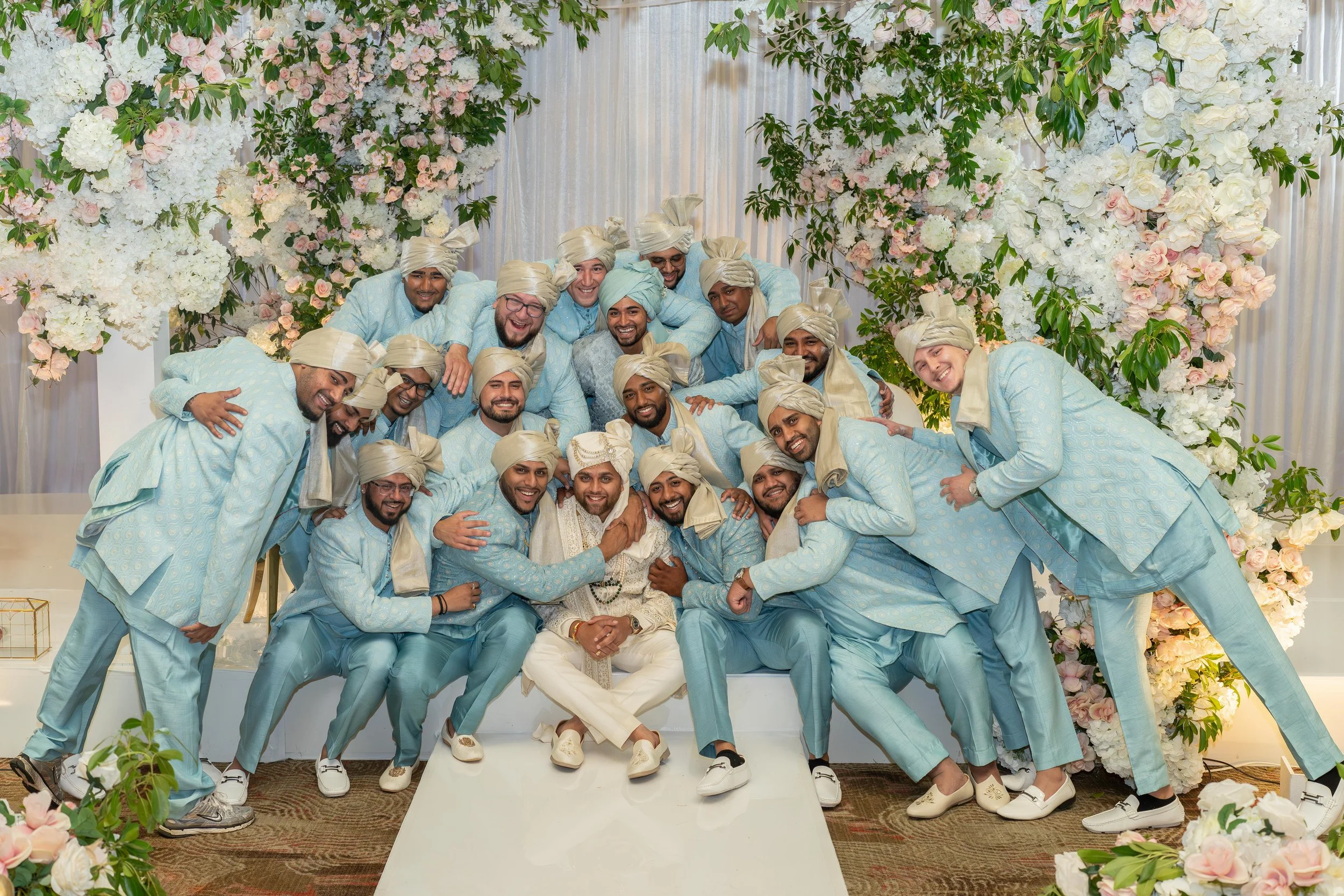 Group of men dressed in light blue traditional Indian attire, wearing beige turbans, gathered together at a celebration, seated and standing in front of a floral backdrop of white and pink flowers with greenery.