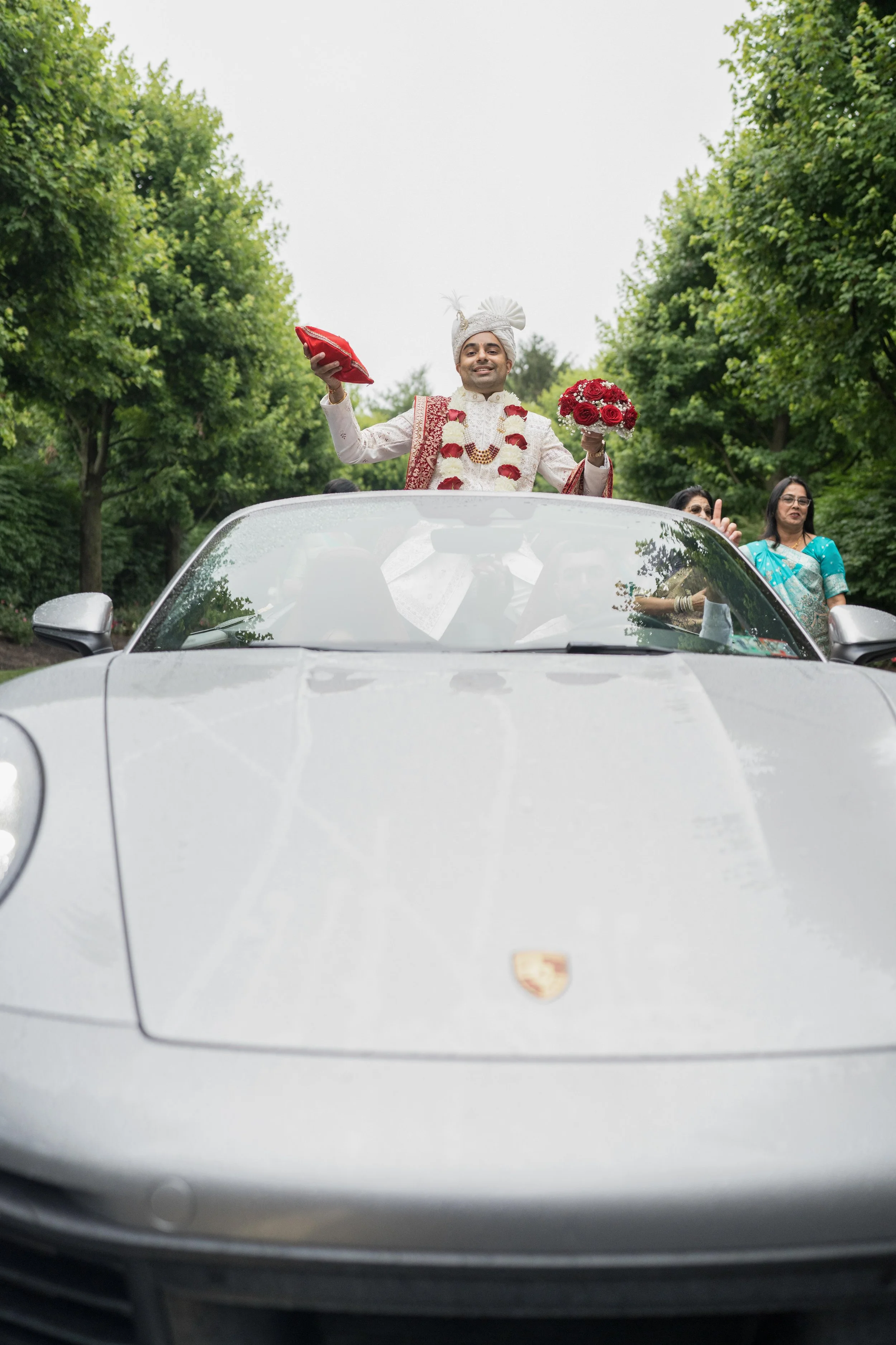 A groom in traditional Indian wedding attire standing through the sunroof of a silver Porsche, holding a bouquet and a pillow, celebrating with friends in a green outdoor setting.