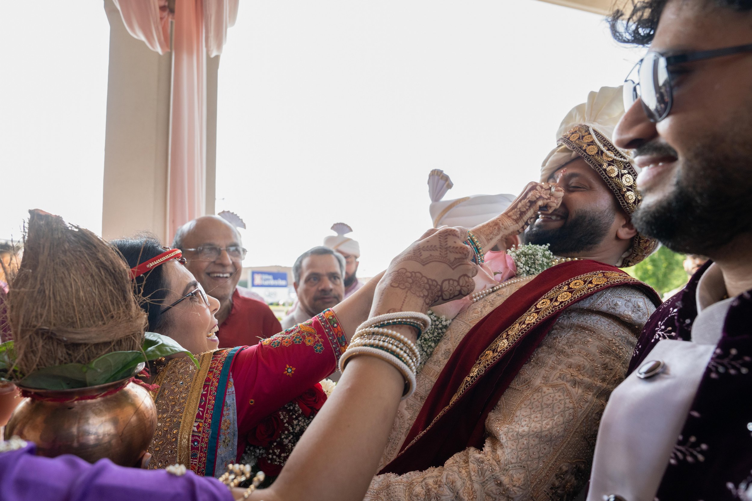 Indian wedding celebration with family and friends, a woman playfully touching the groom's nose, traditional attire, colorful garments, joyous atmosphere.