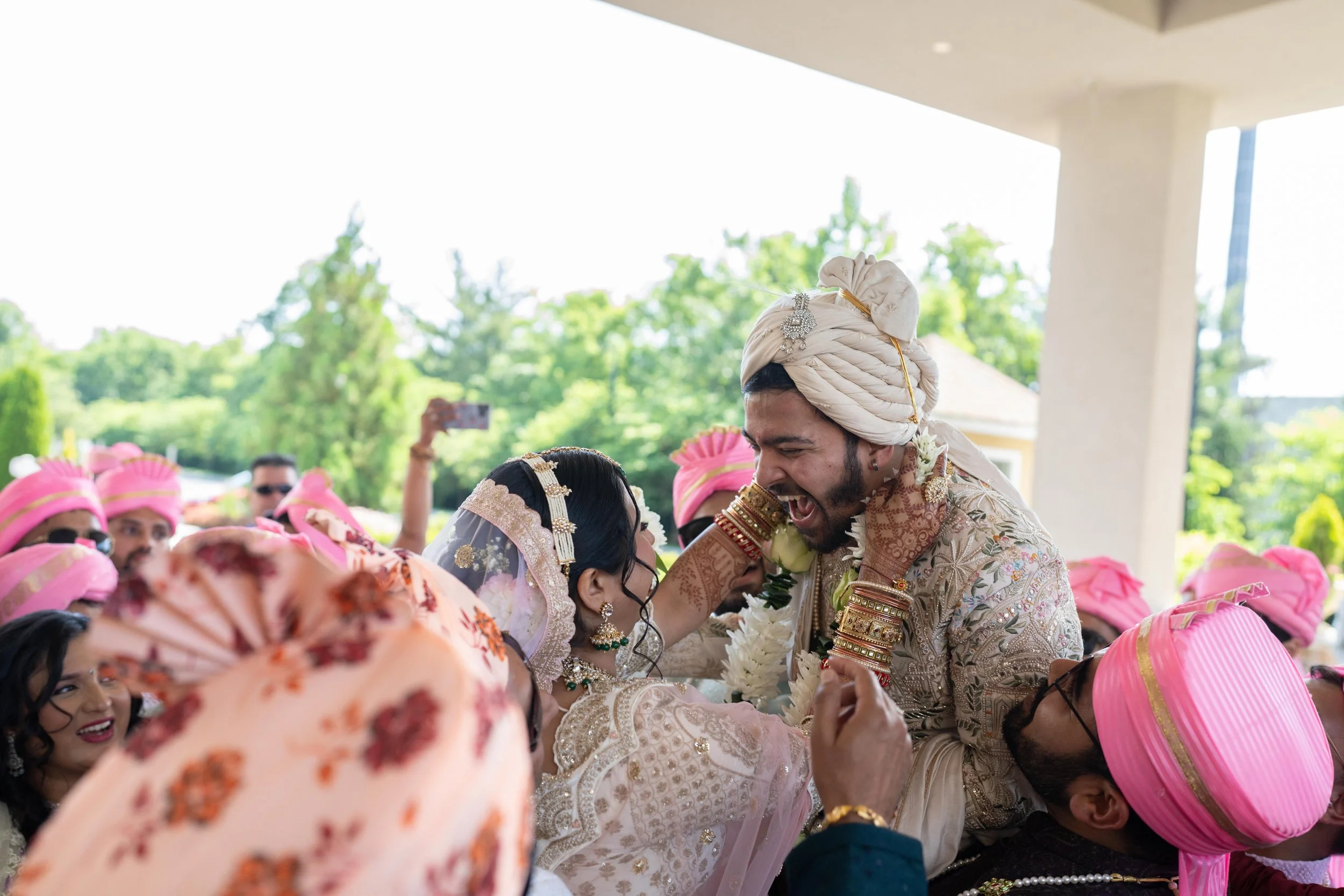 Indian bride and groom celebrating during a wedding ceremony, surrounded by guests wearing traditional attire and pink turbans, outdoors with green trees in the background.