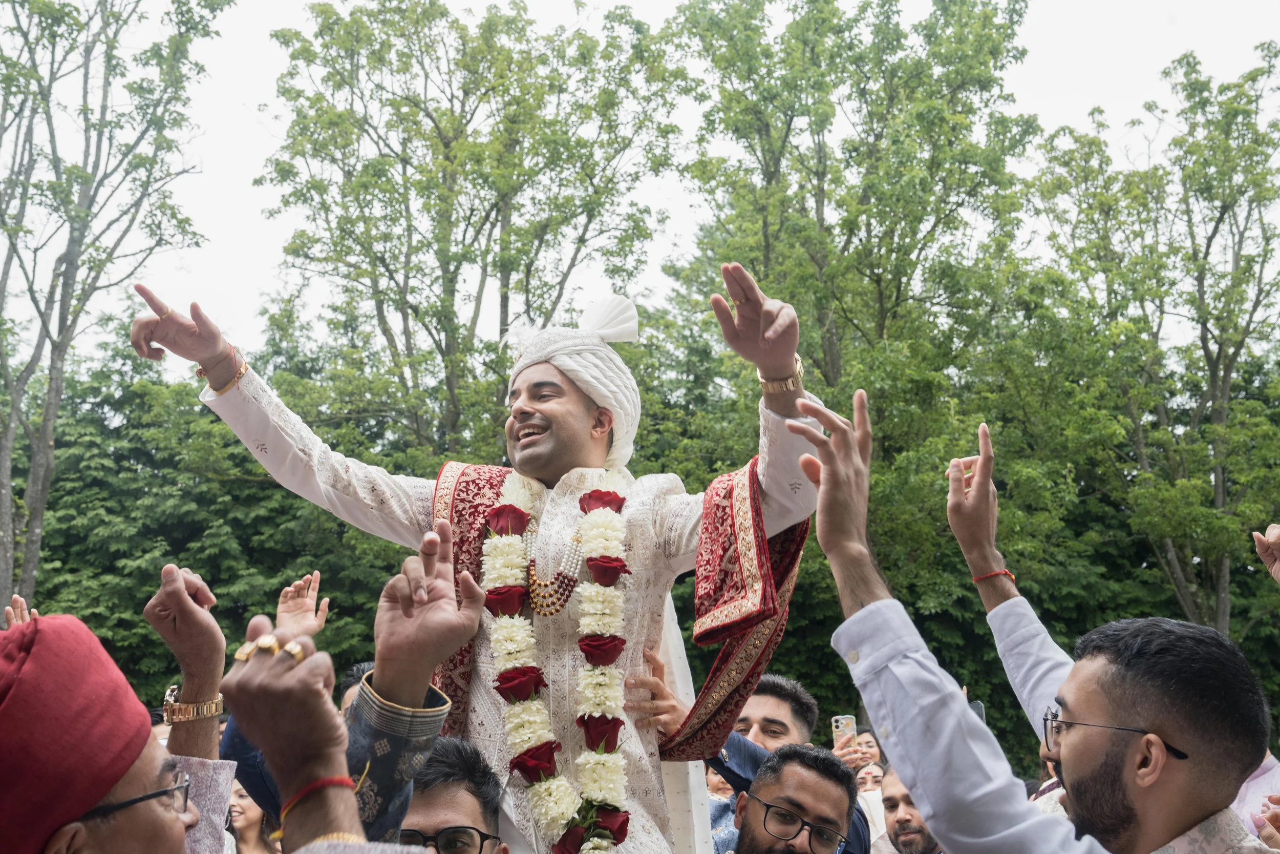 Groom in traditional Indian wedding attire being lifted by a crowd, wearing a garland of flowers, surrounded by people celebrating outdoors with trees in the background.
