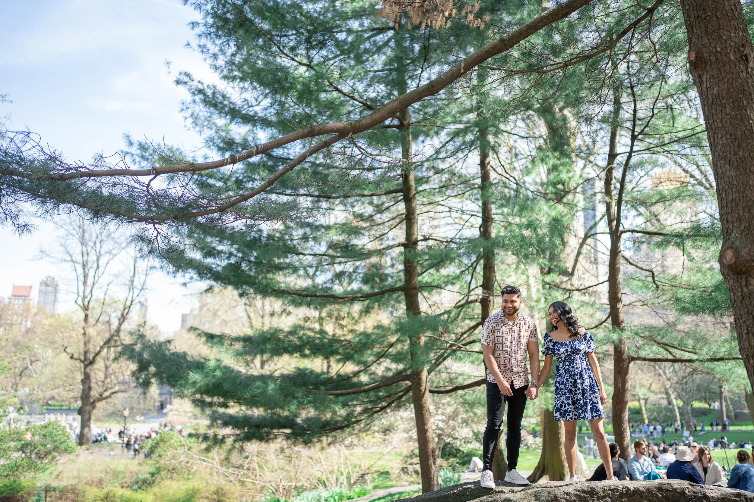 A smiling groom and bride holding hands, standing on a large rock in a central park with tall trees and a crowd of people in the background on a sunny day.