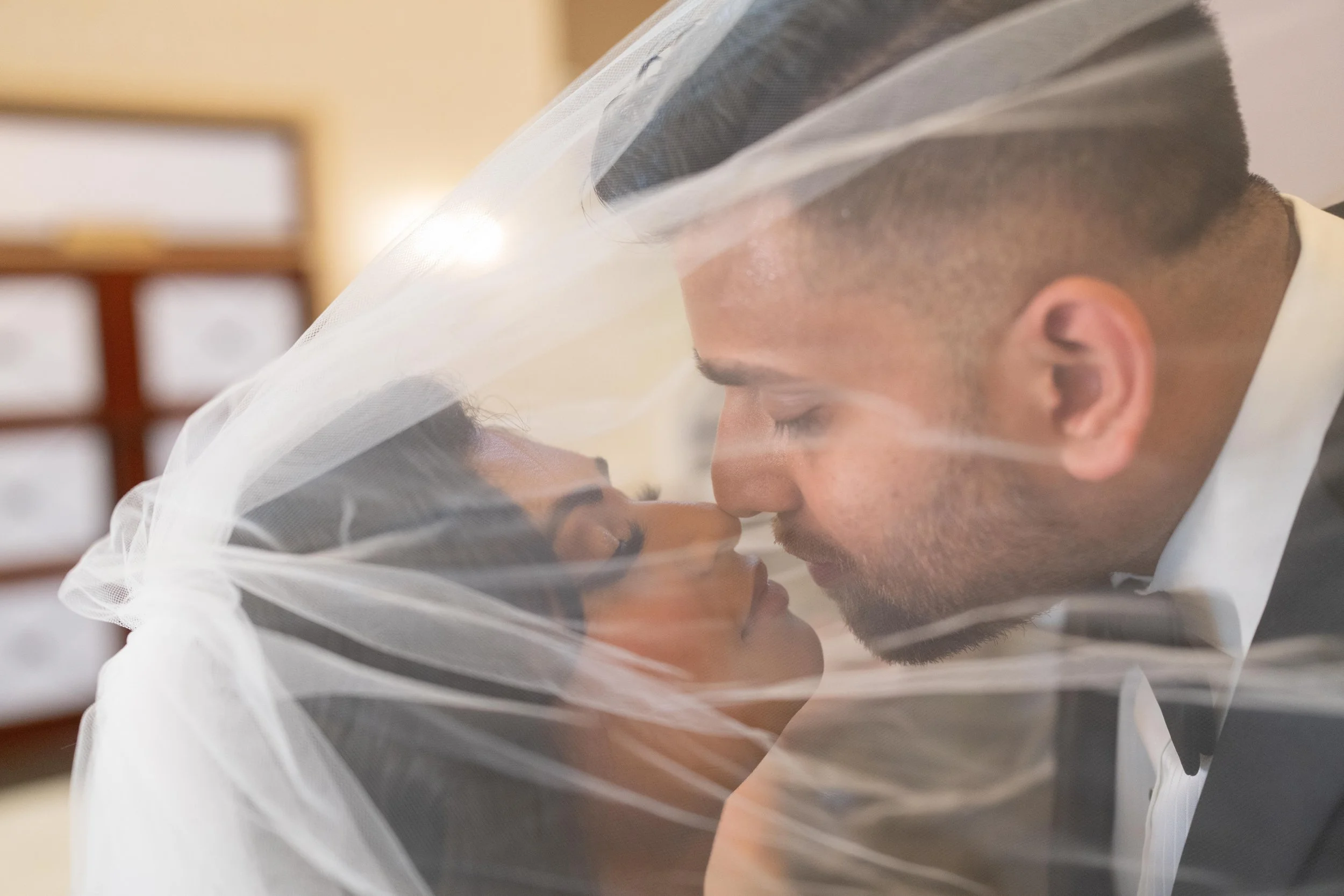 A bride and groom leaning in close together under a veil for a wedding photo.
