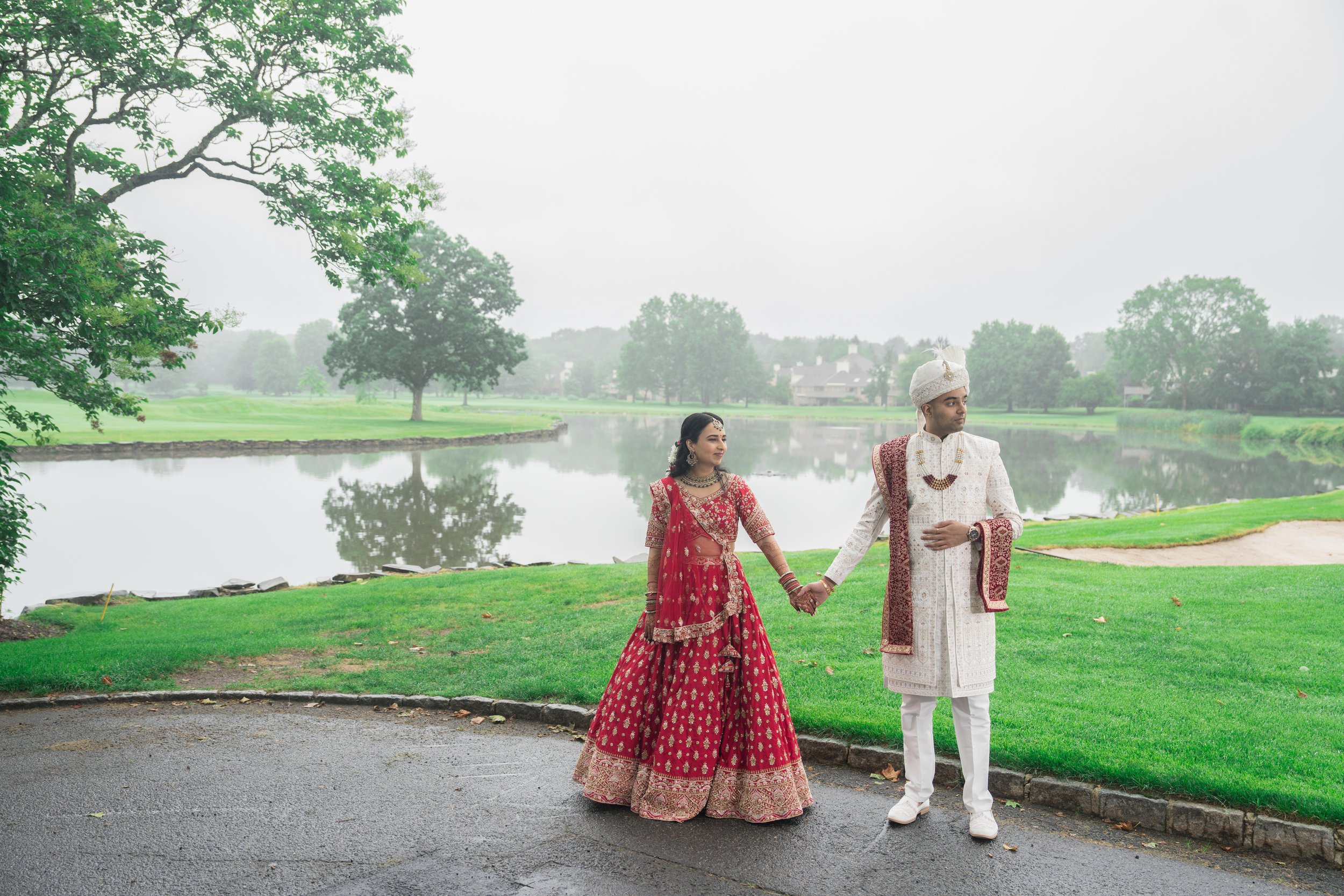 A couple dressed in traditional Indian wedding attire holding hands near a lake with trees and houses in the background.