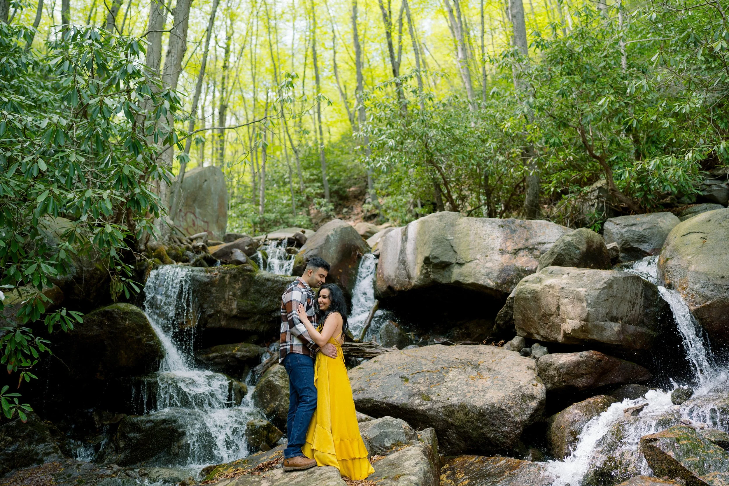 A couple standing on rocks near a small waterfall in a lush green forest, embracing each other and smiling.