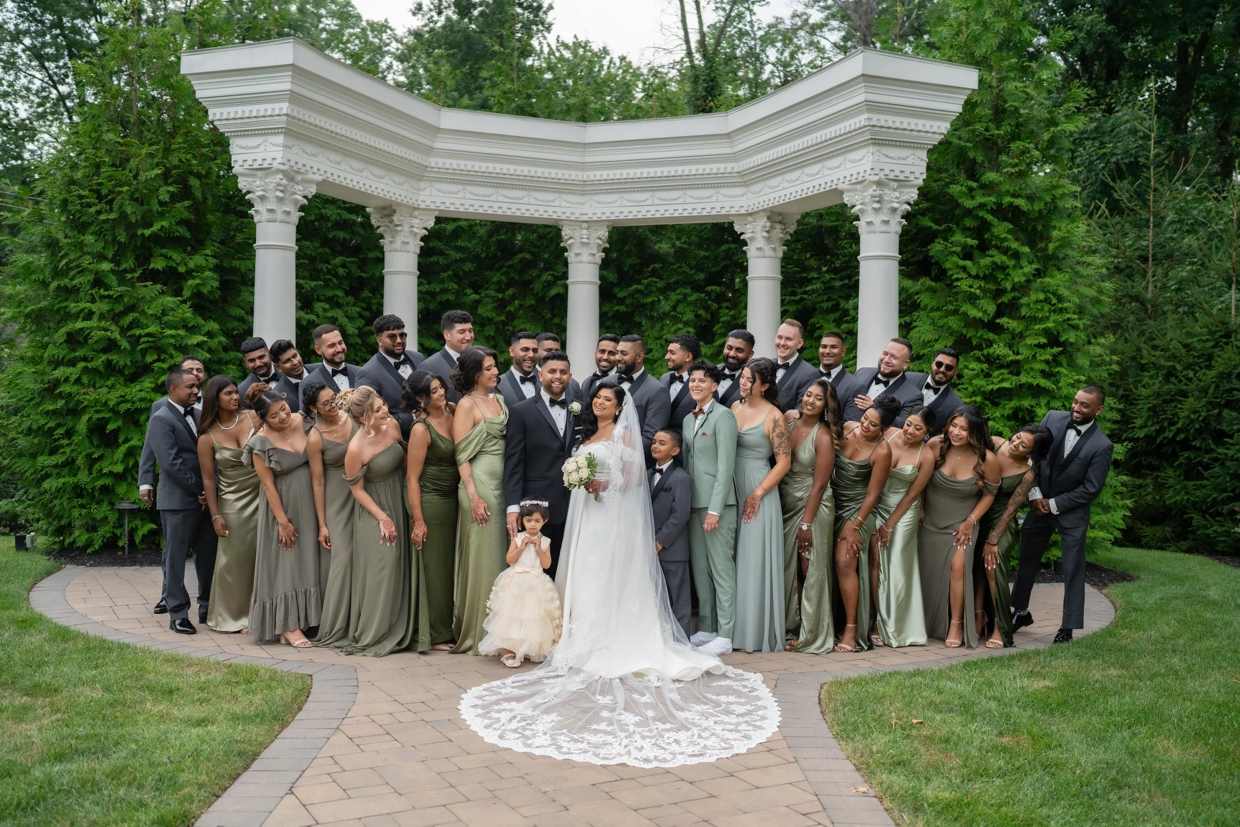 A large group of wedding guests, including the bride and groom, standing outdoors on a brick pathway under a white architectural pergola surrounded by green trees.