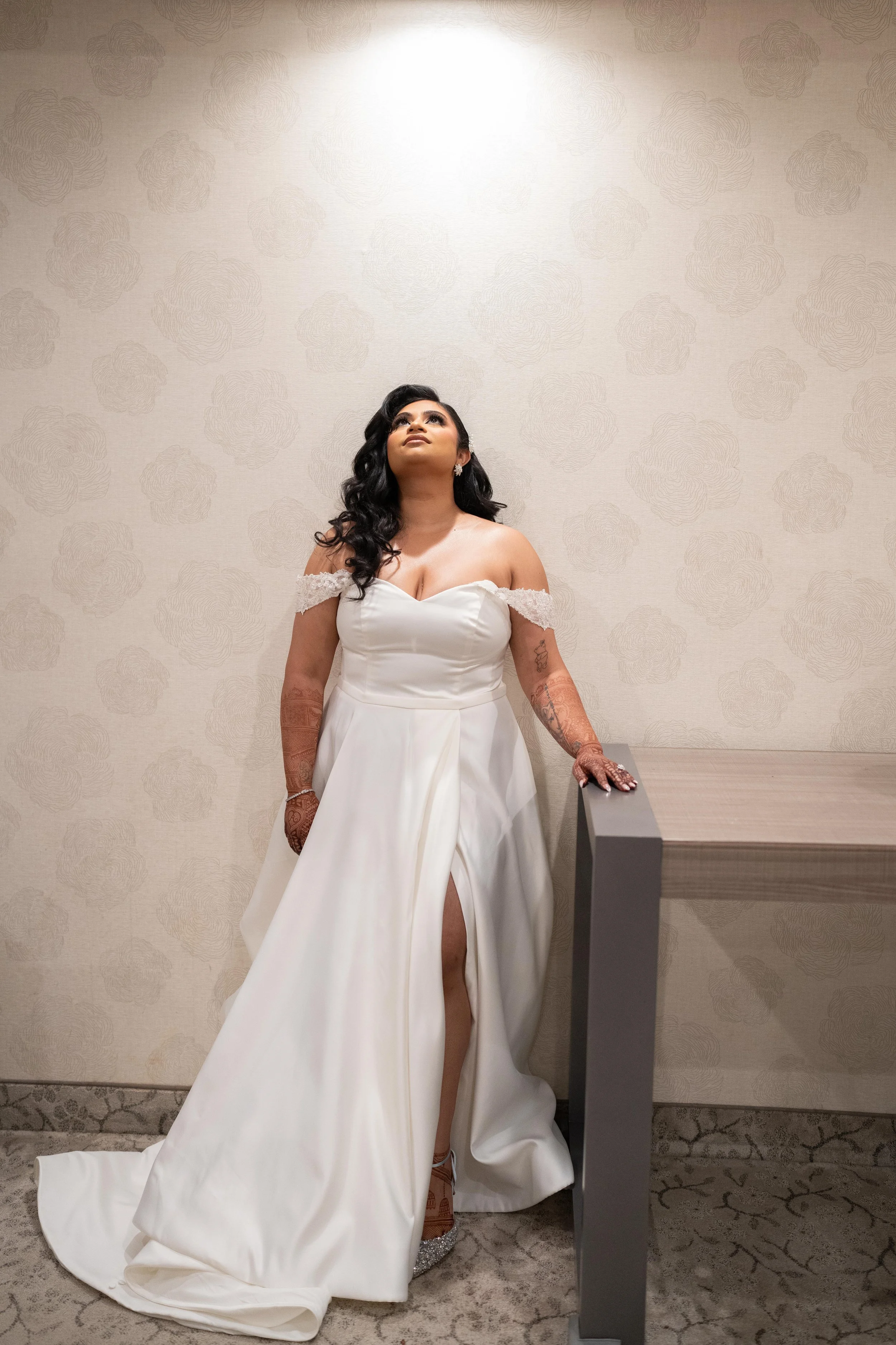 A bride in a white wedding gown with off-shoulder lace details, standing beside a table, looking upwards, with black wavy hair and henna tattoos on her arms, in front of a patterned wall.