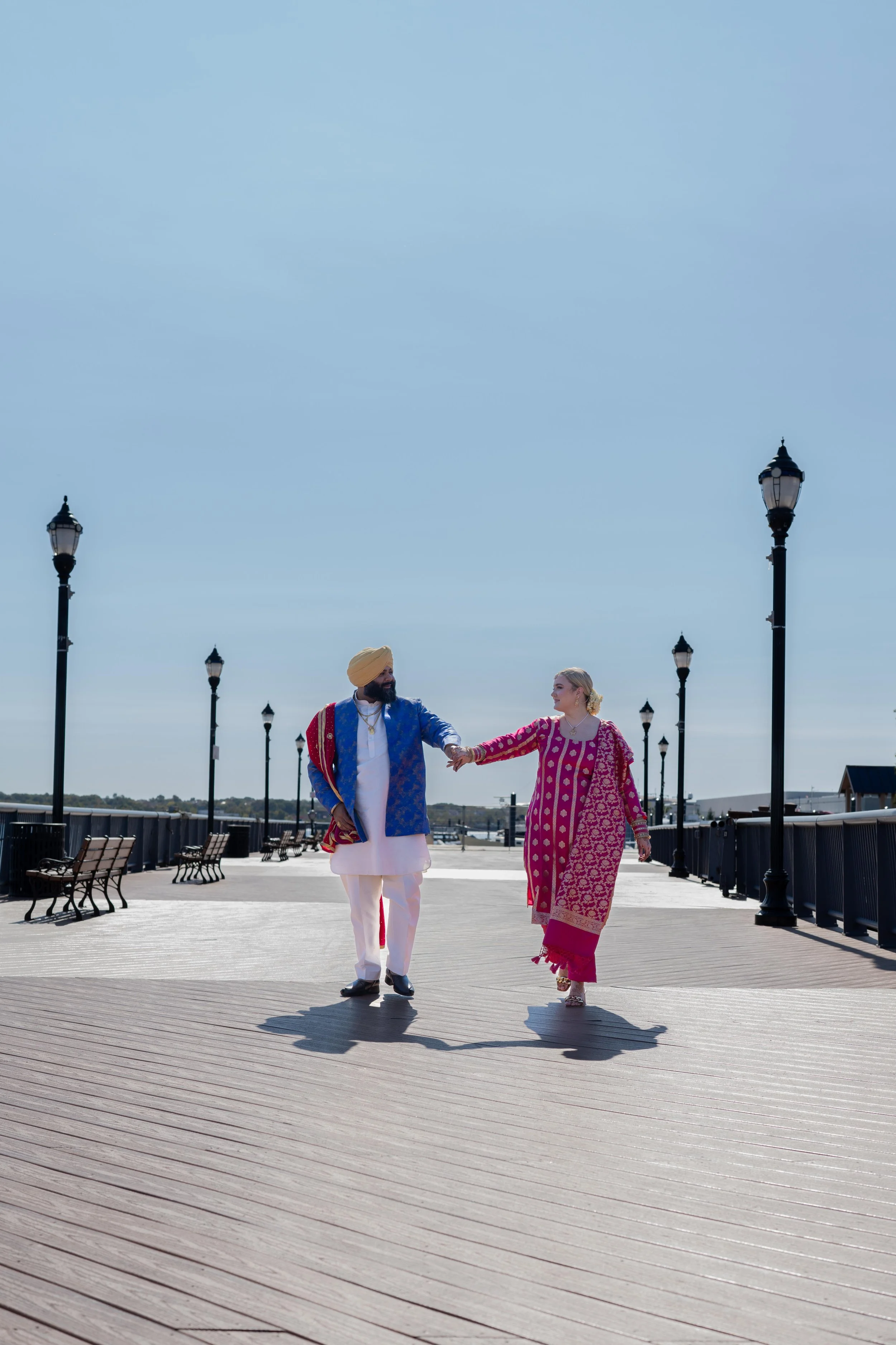 A bride and groom dressed in traditional Indian clothing are walking hand-in-hand on a pier, with lampposts and benches visible along the path and a clear blue sky overhead.