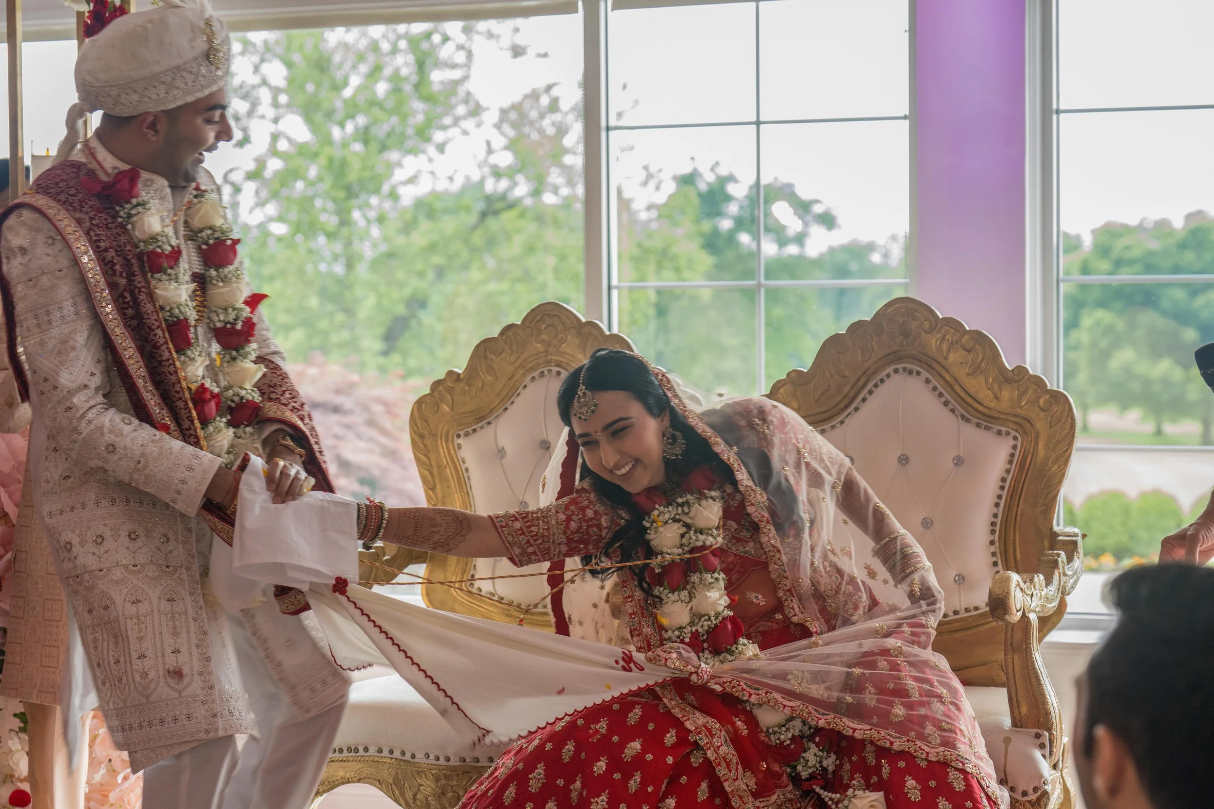 A bride and groom during a traditional Indian wedding ceremony. The bride, dressed in a red saree with gold embroidery, is sitting on a decorated throne, smiling and reaching out. The groom, standing next to her, is wearing a white and gold outfit wi