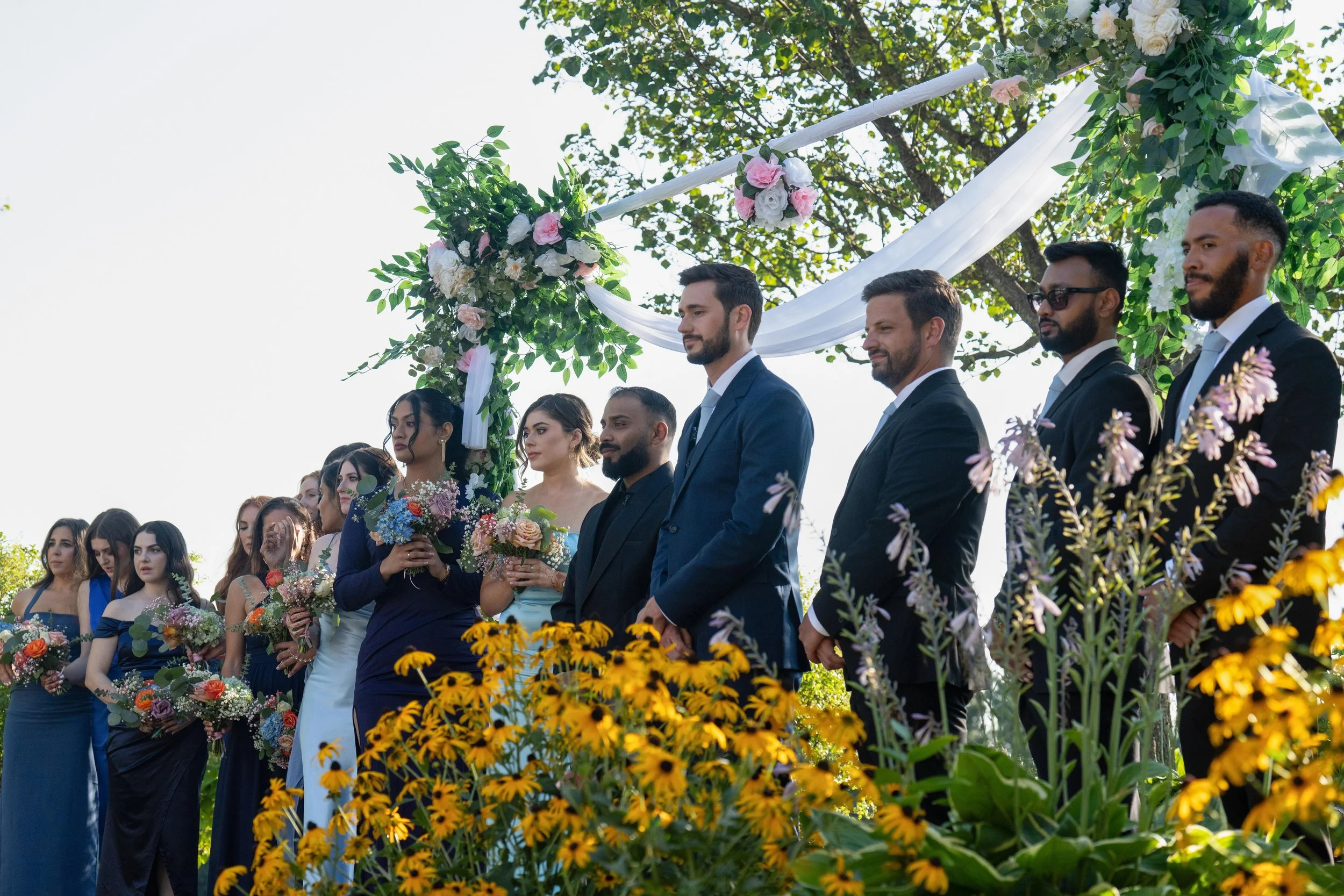 A wedding ceremony outdoors with bridesmaids in blue dresses and groomsmen in suits standing under a floral arch, surrounded by yellow flowers and greenery.