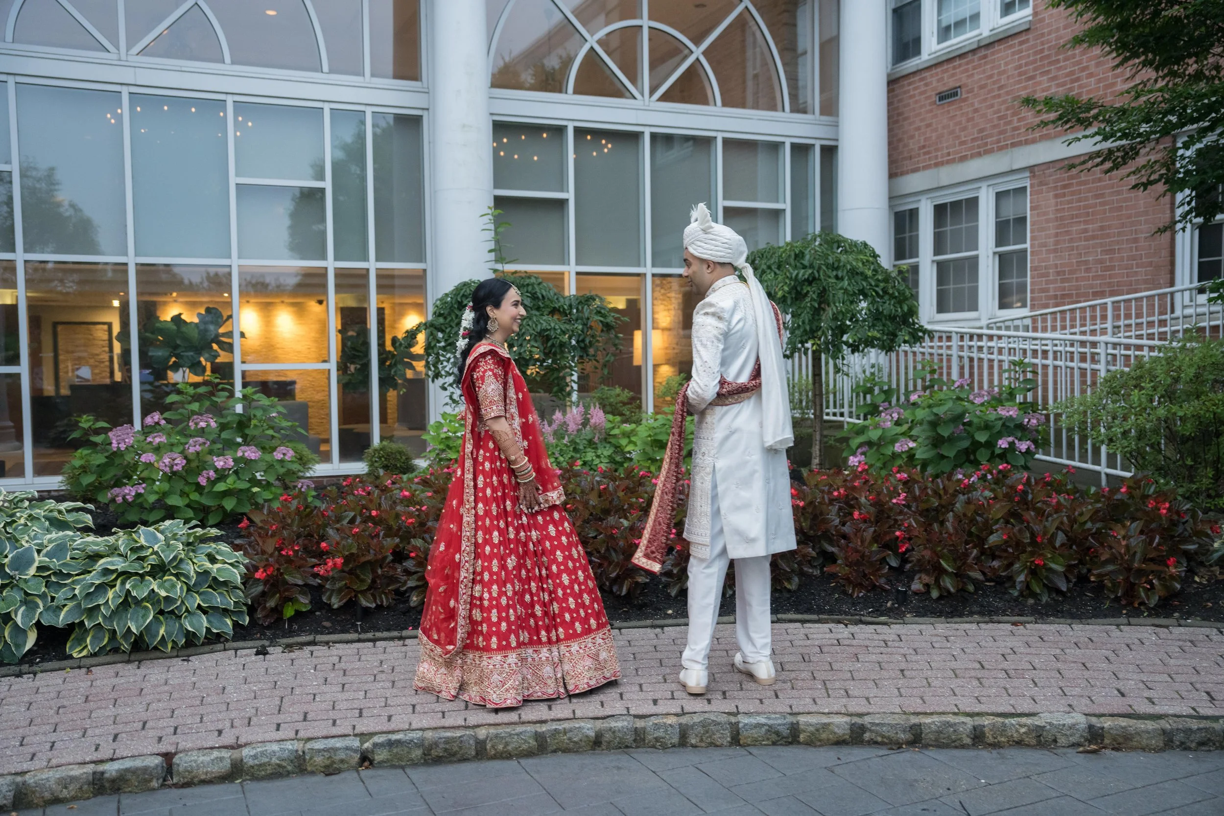A couple dressed in traditional Indian wedding attire standing outdoors on a brick pathway, facing each other and smiling, with a glass building and garden with pink and purple flowers behind them.