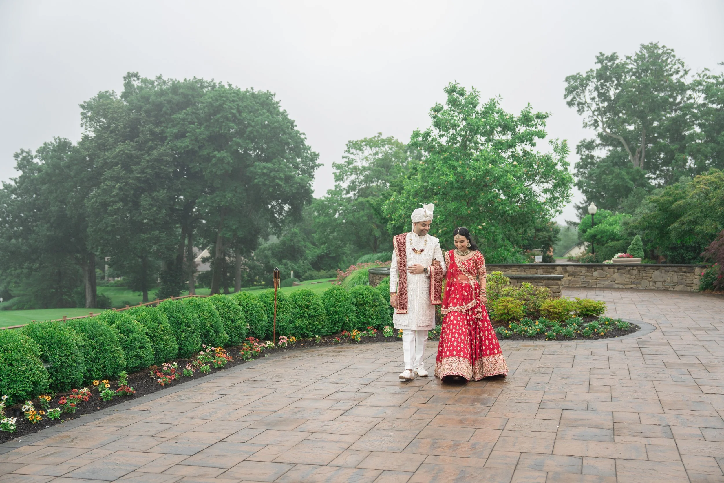 A traditionally dressed Indian couple walking together outdoors on a paved pathway surrounded by lush greenery and flowers.