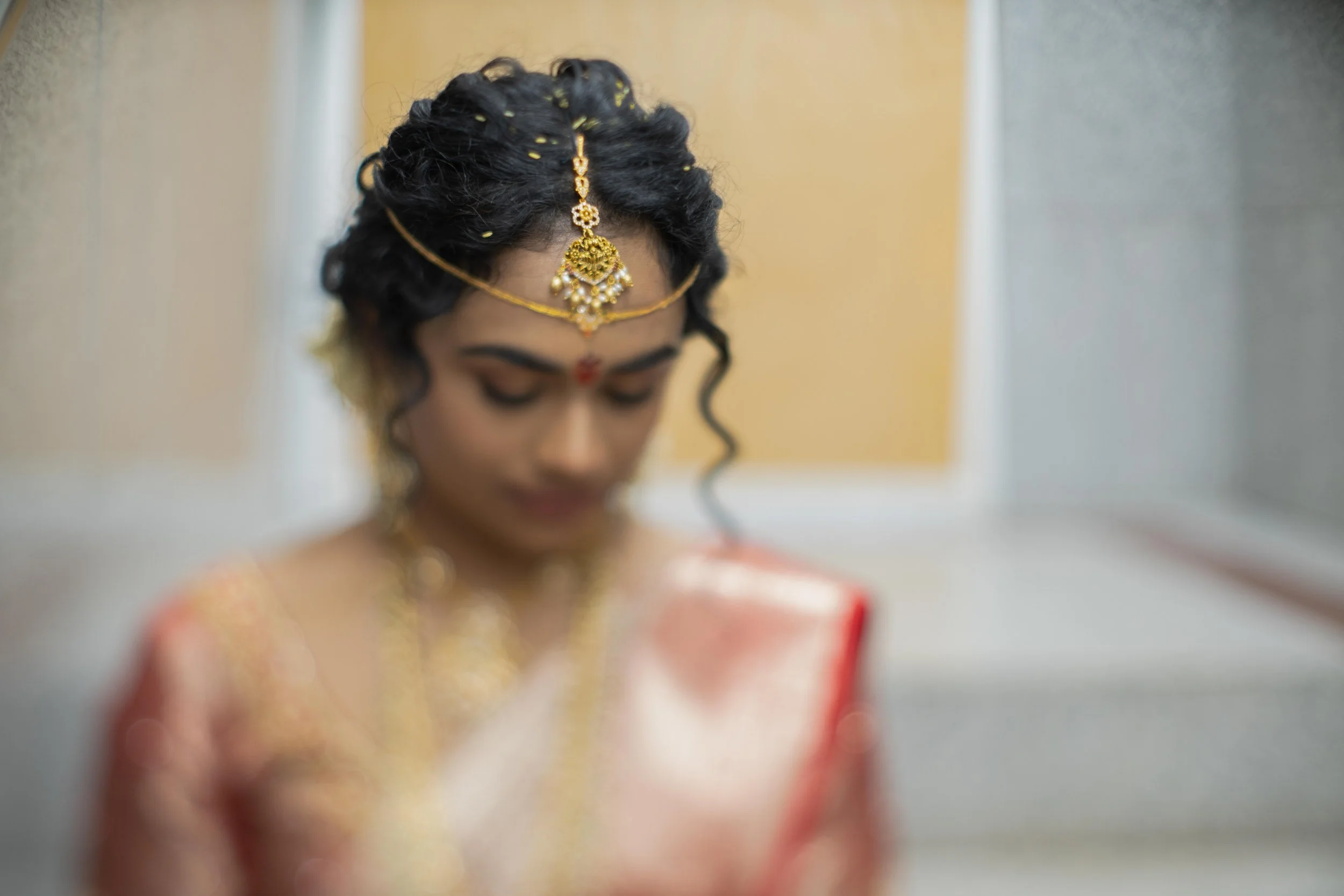 A bride dressed in traditional Indian wedding attire, including jewelry and a red garment, is looking down.