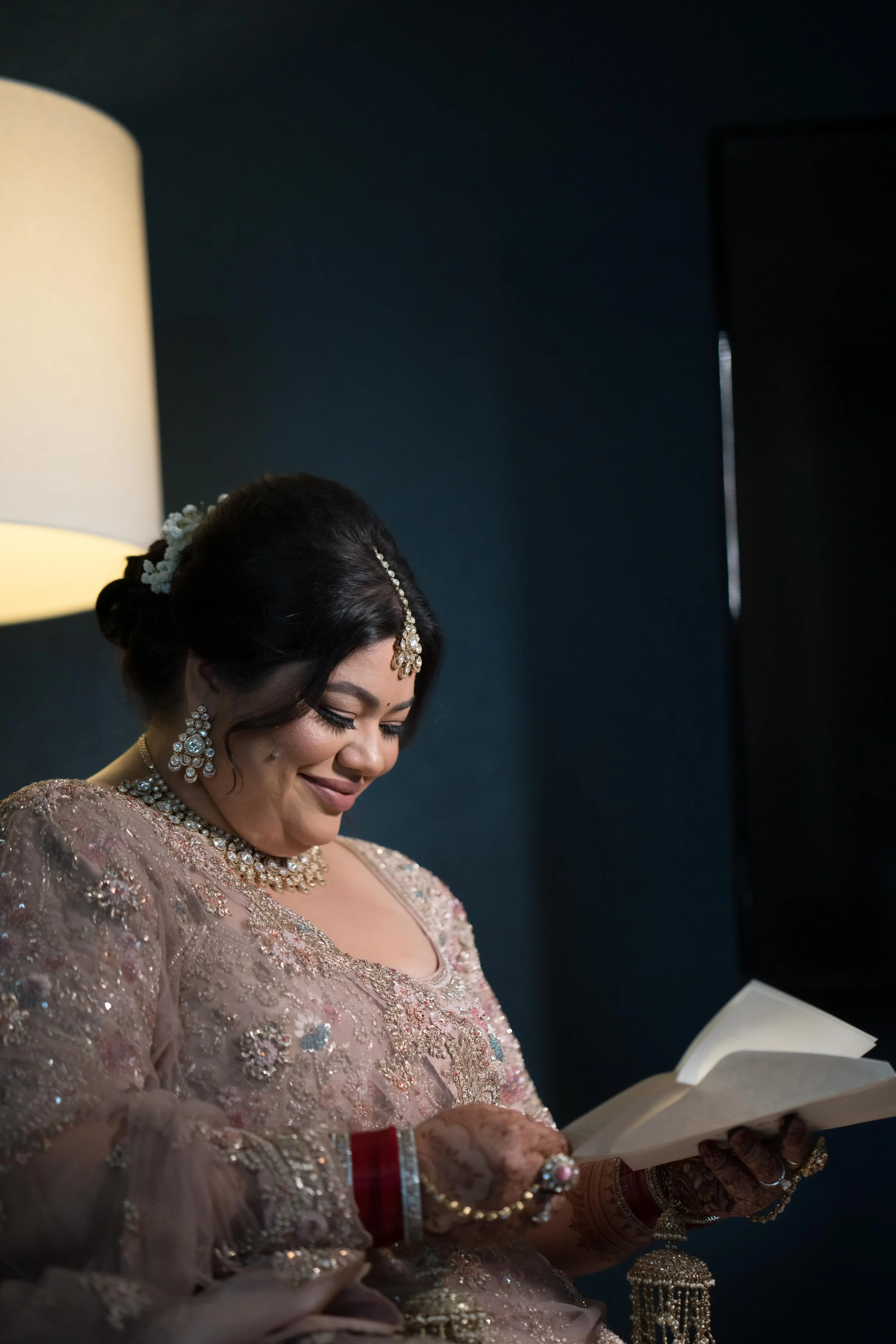 A bride dressed in traditional Indian bridal attire, with jewelry and henna on her hands, reading a book and smiling.