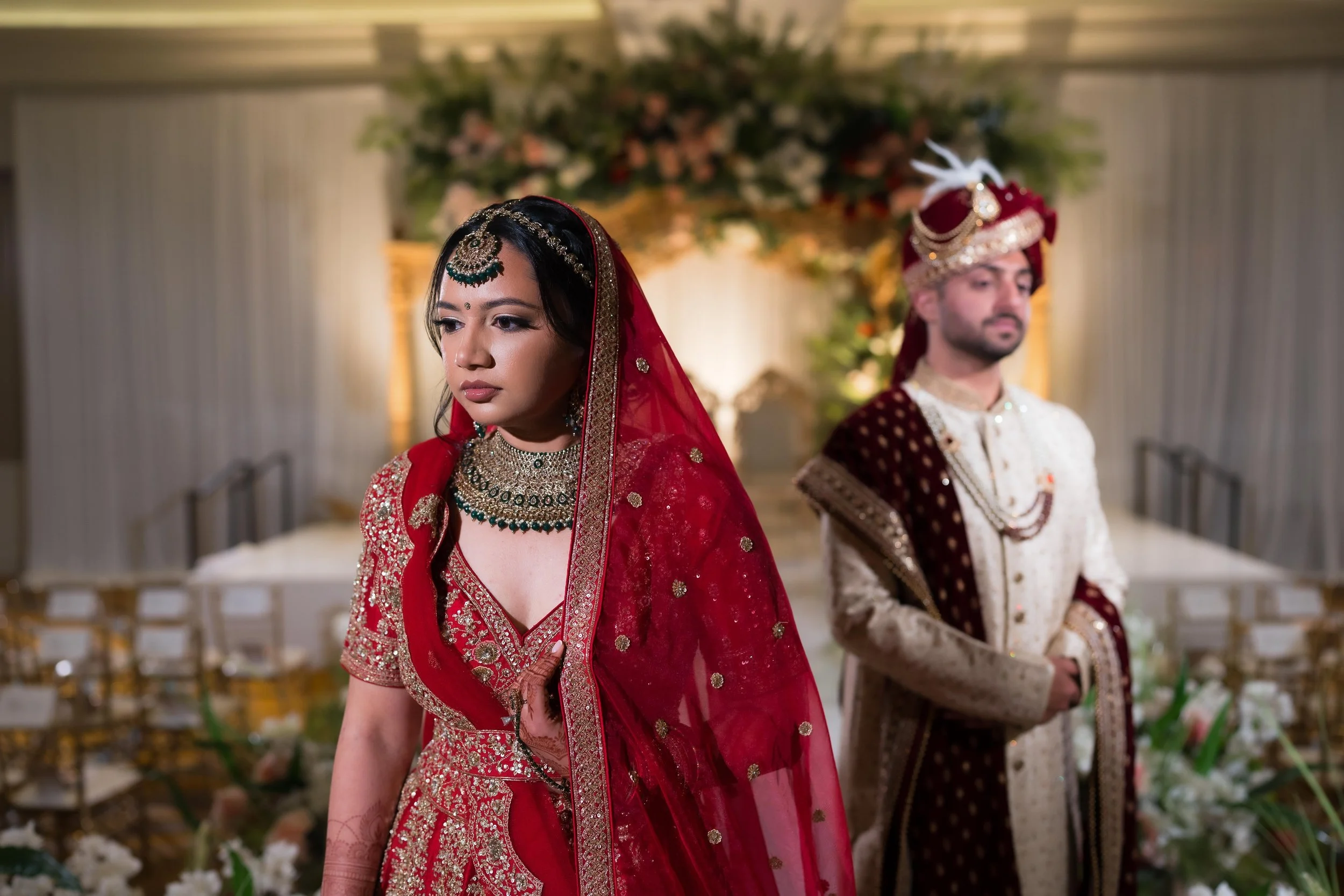 Indian bride and groom in traditional wedding attire, standing separately during a wedding ceremony, with floral decorations in the background.