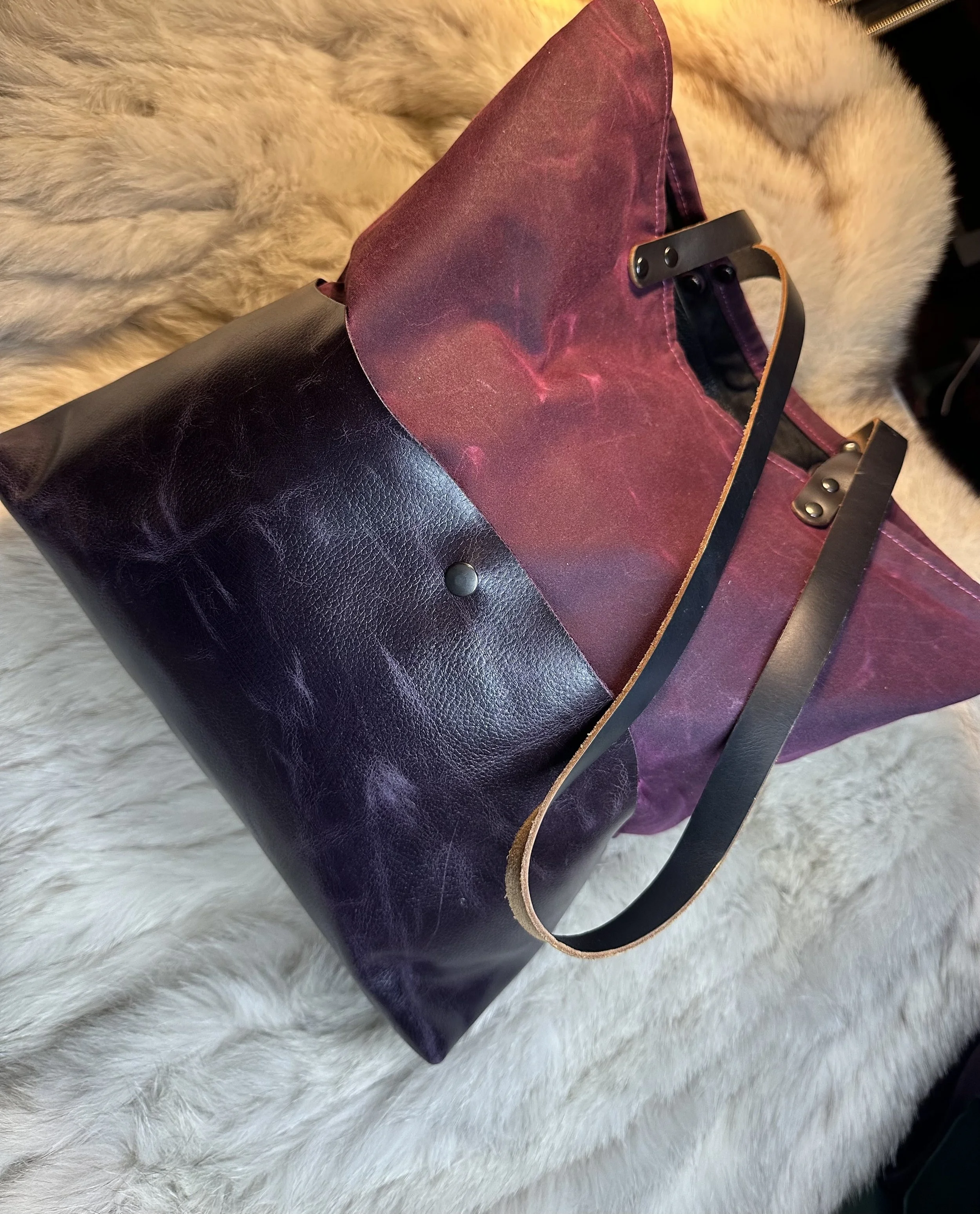 Close-up of a black leather handbag resting on a white fluffy surface, with a purple fabric section and a metal handle.