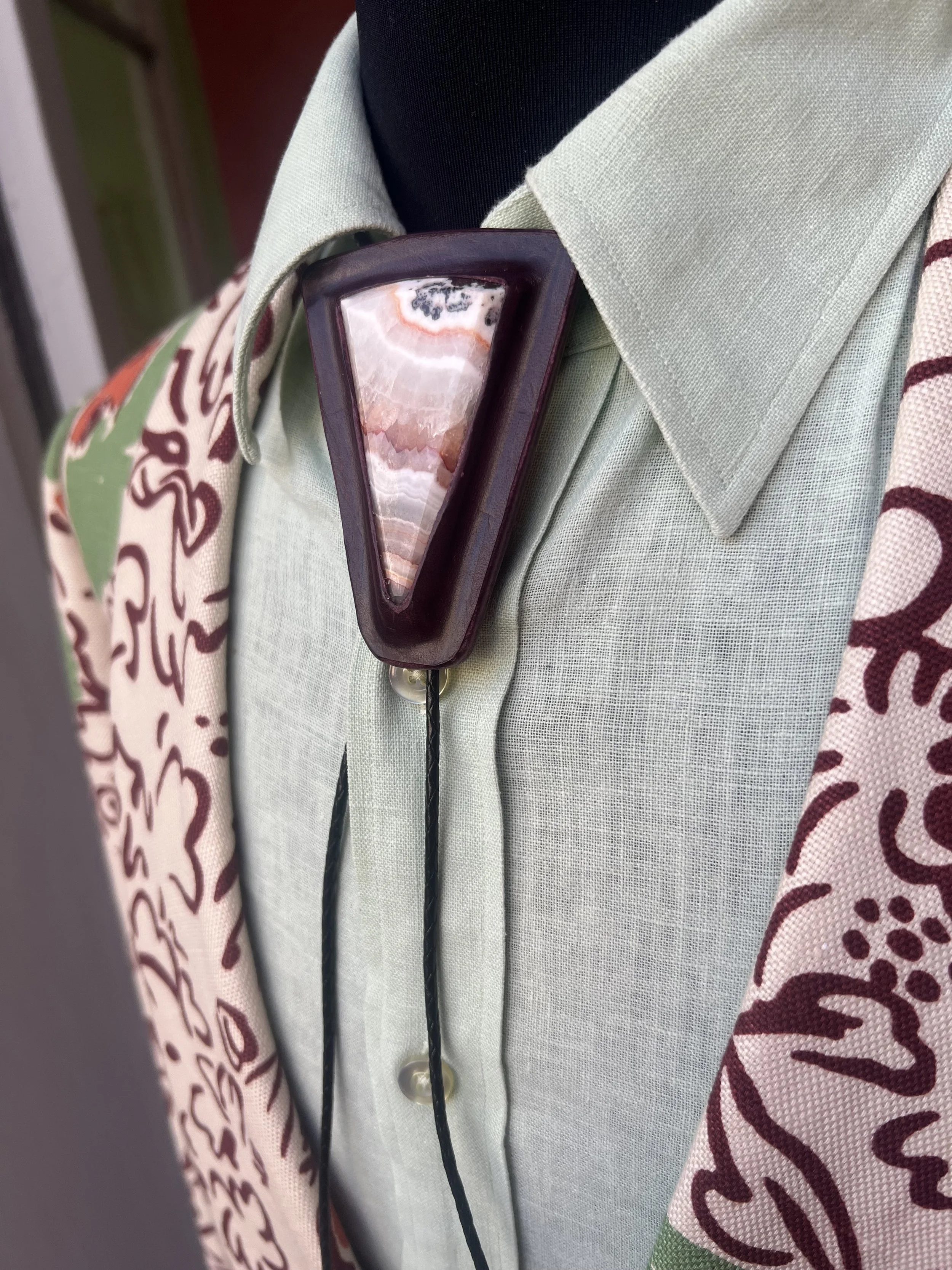 Close-up of a person's shoulder wearing a light green blazer, a black shirt, and a scarf with a floral pattern. A large, triangular onyx and marble brooch is pinned to the blazer.