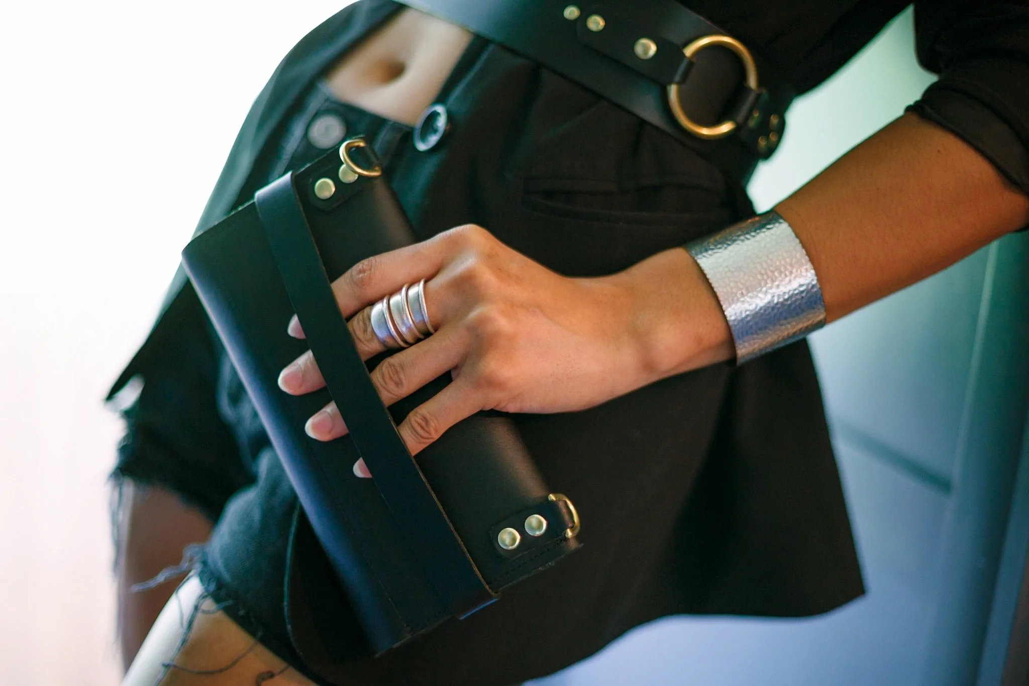 Close-up of a person wearing a black outfit with metallic accessories, including a wide metallic bracelet, a black belt with gold rings, and silver rings, holding a black bag with gold stud details.