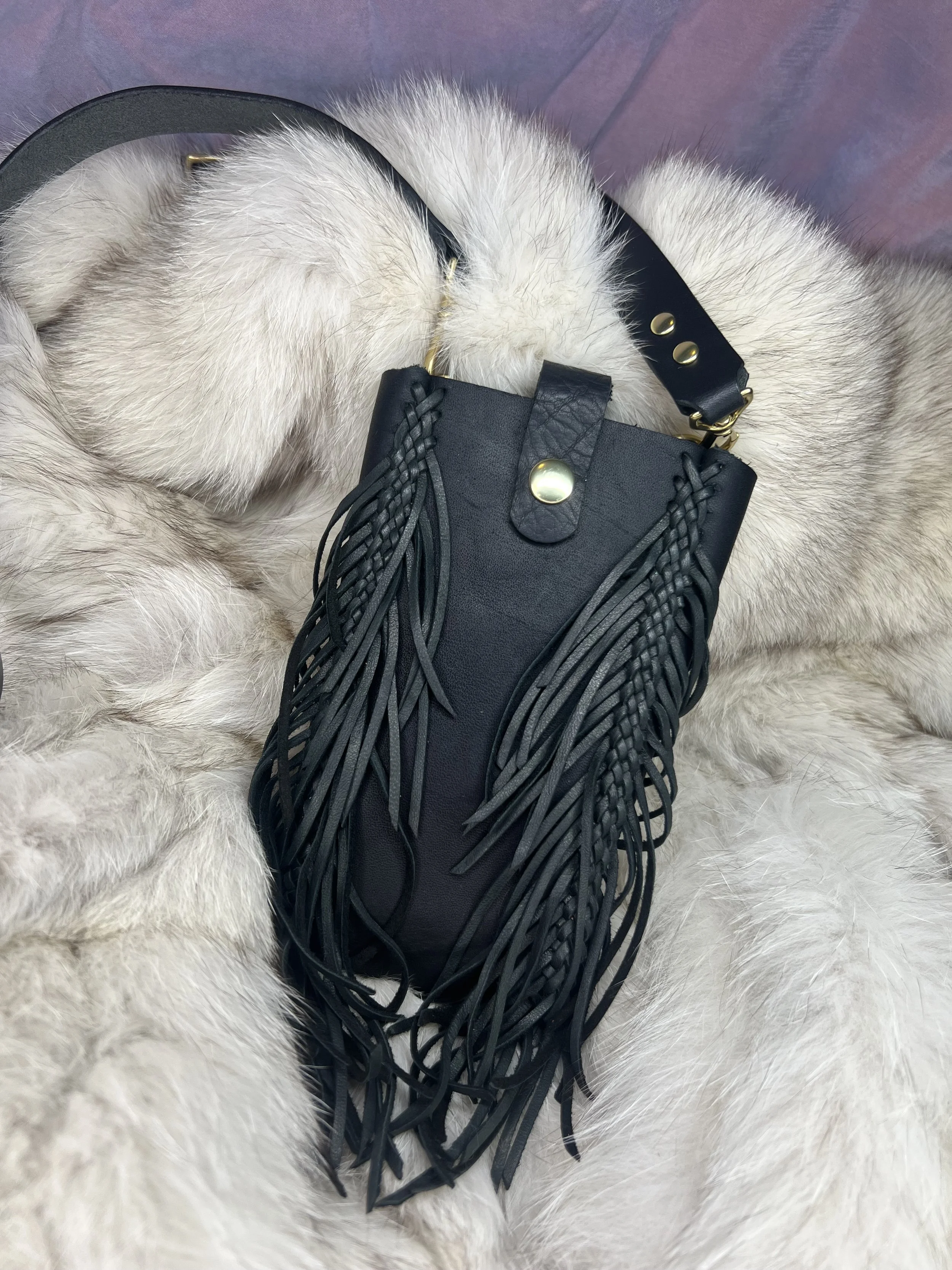 A black handbag with fringe details resting on a fluffy white fur surface.