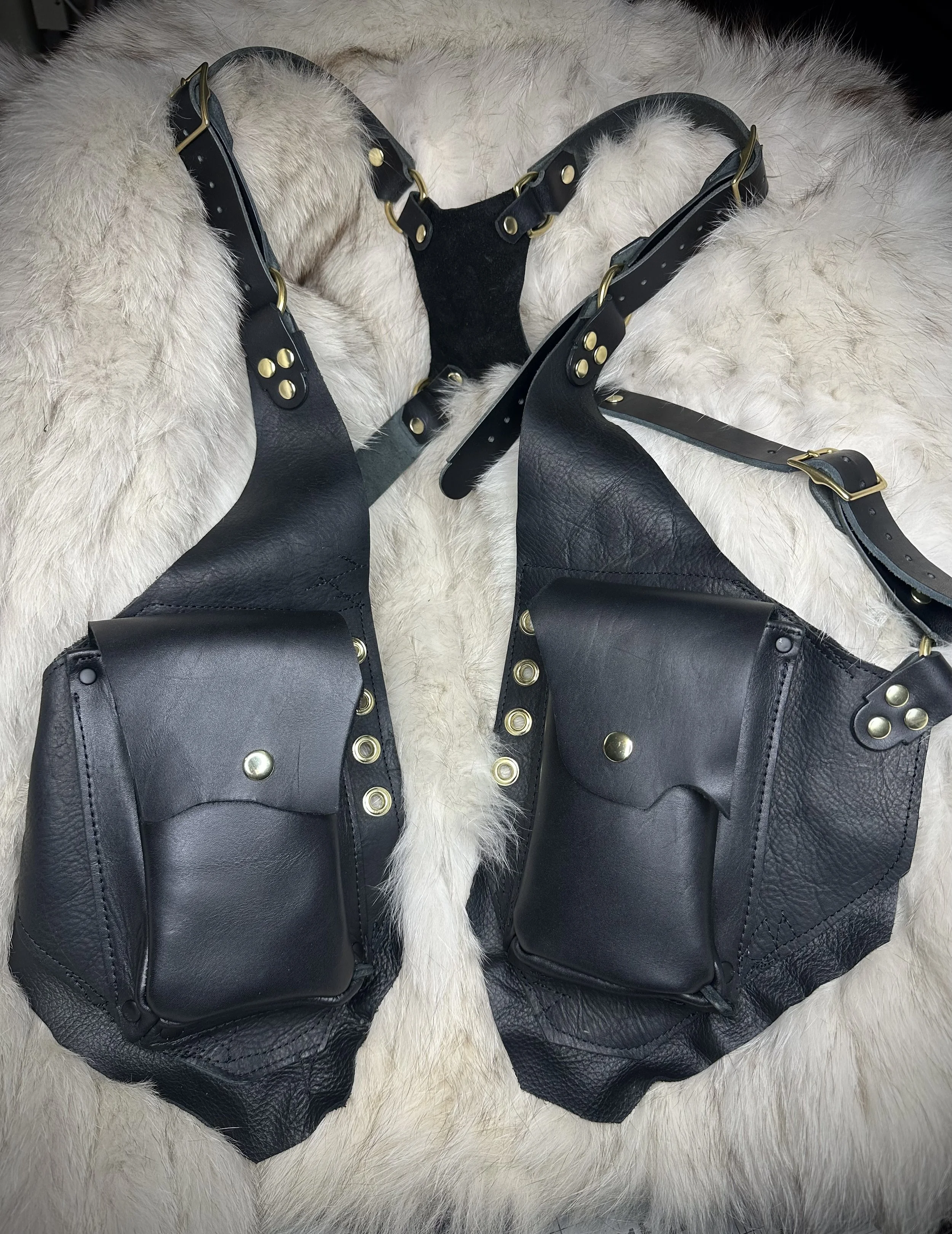 A pair of black leather harnesses with gold-colored hardware, placed on a fluffy white surface.