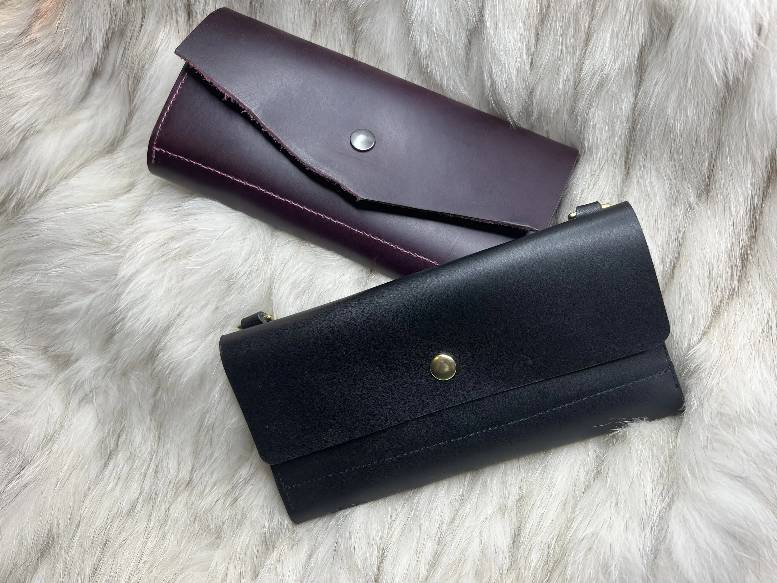 A dark purple leather wallet purse with clean stitching and a silver snap button, placed on a white furry surface. Colorful customizable wallets that are multi wear, & can be worn as a purse with detachable strap. 