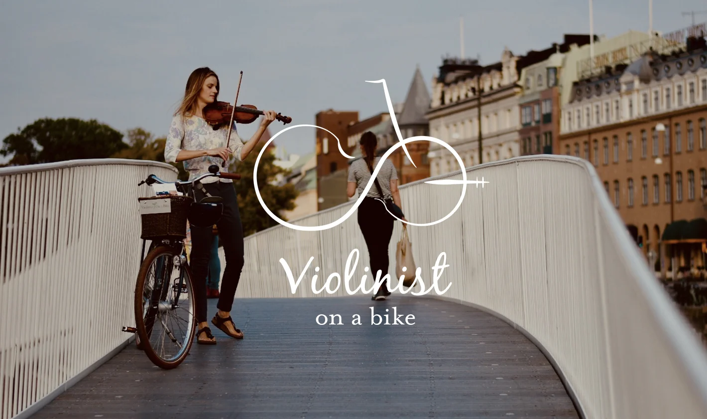To kvinder går på en bro i en by, en spiller violin og har en cykel, mens den anden går væk. Tekst over billedet siger 'Violinist on a bike'.