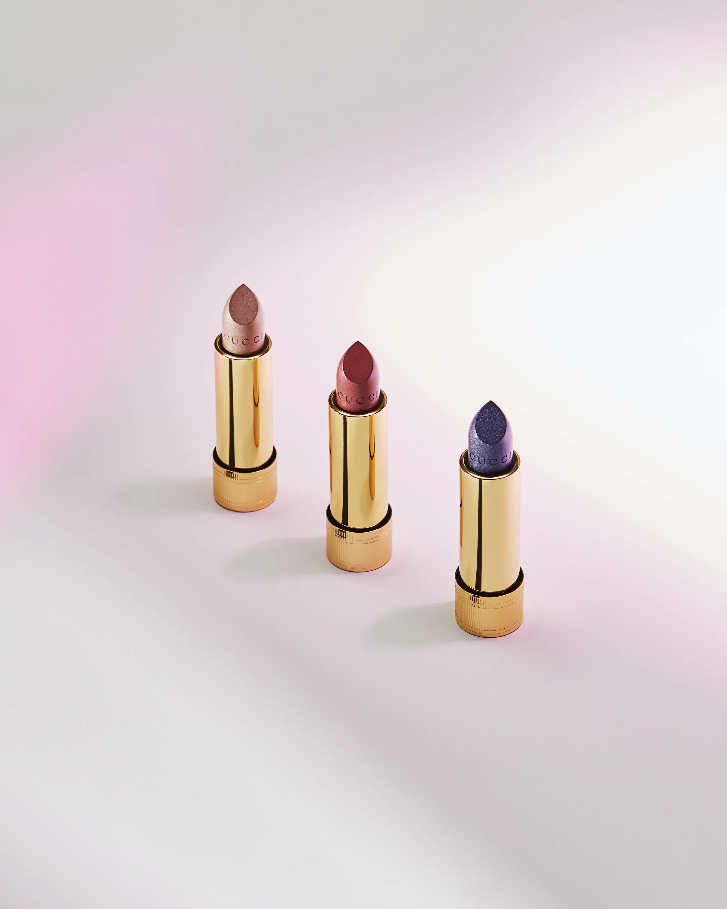 Three Gucci lipsticks with gold tubes, arranged in a row on a white surface, with pink, red, and purple shades.
