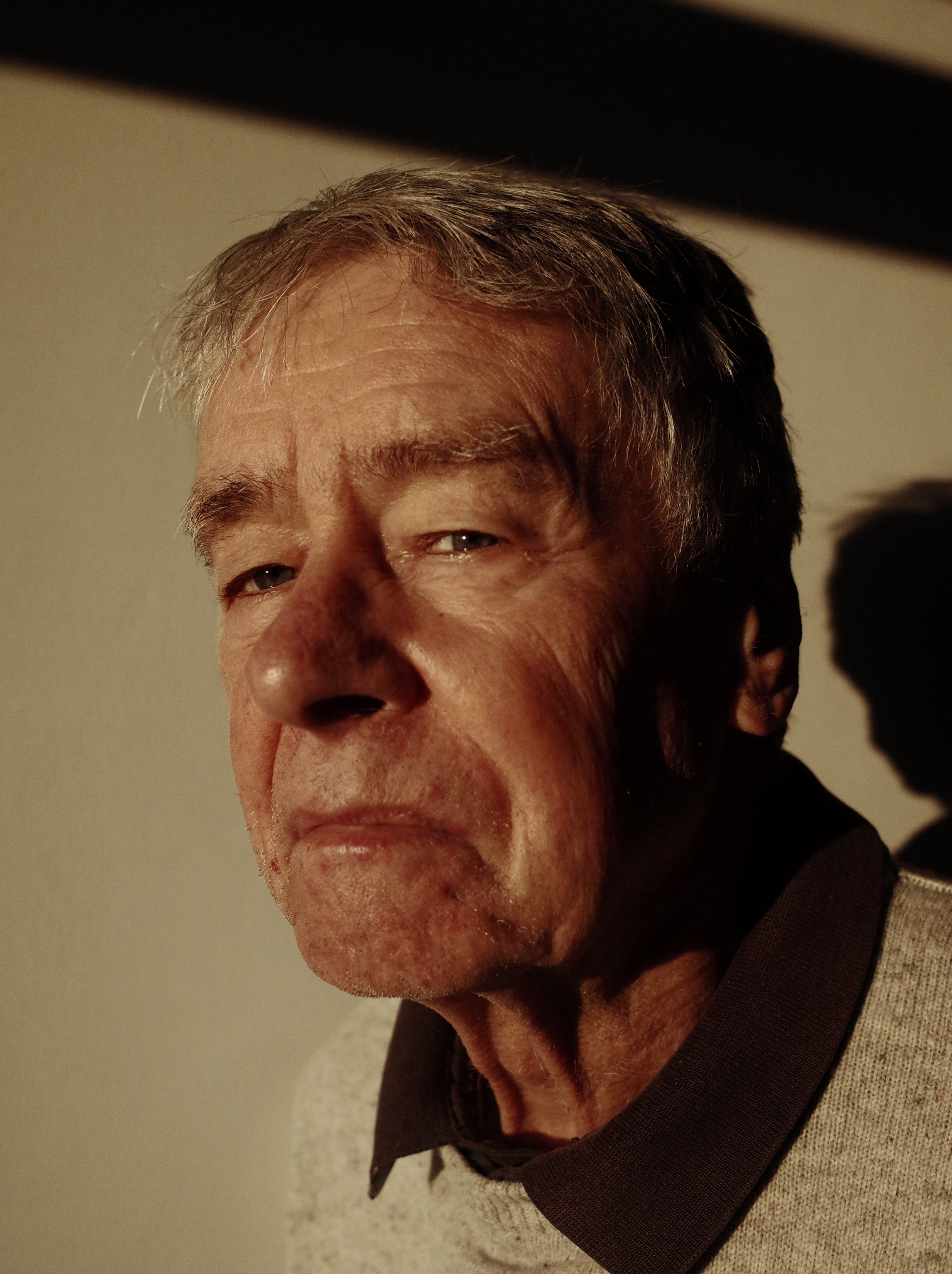 Close-up of an older man with gray hair, looking at the camera with a slight squint, against a plain background.