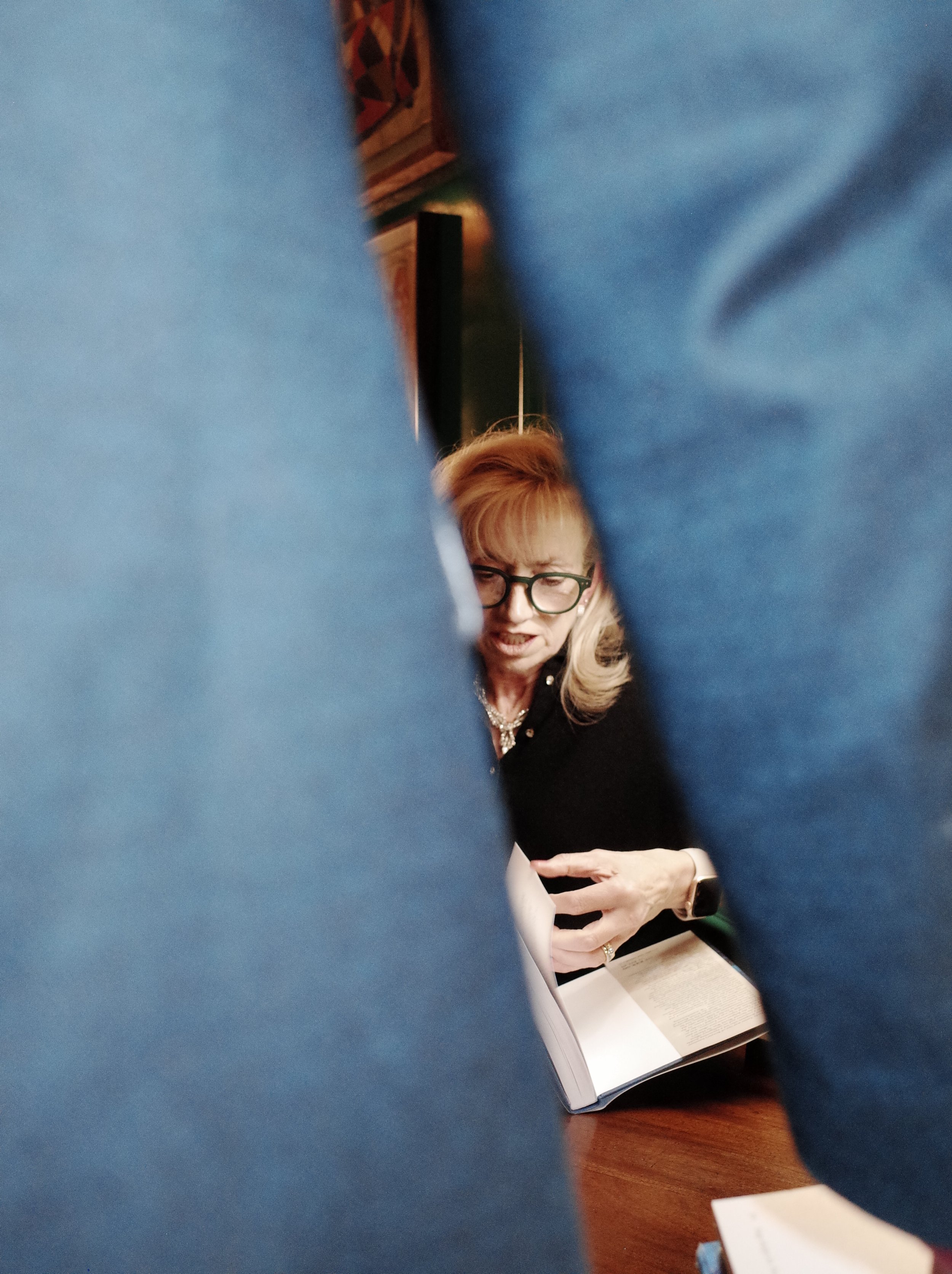 A woman with blonde hair, glasses, and a black outfit sitting at a table, reading a book, and making a facial expression, seen through a gap between two people wearing blue clothing.