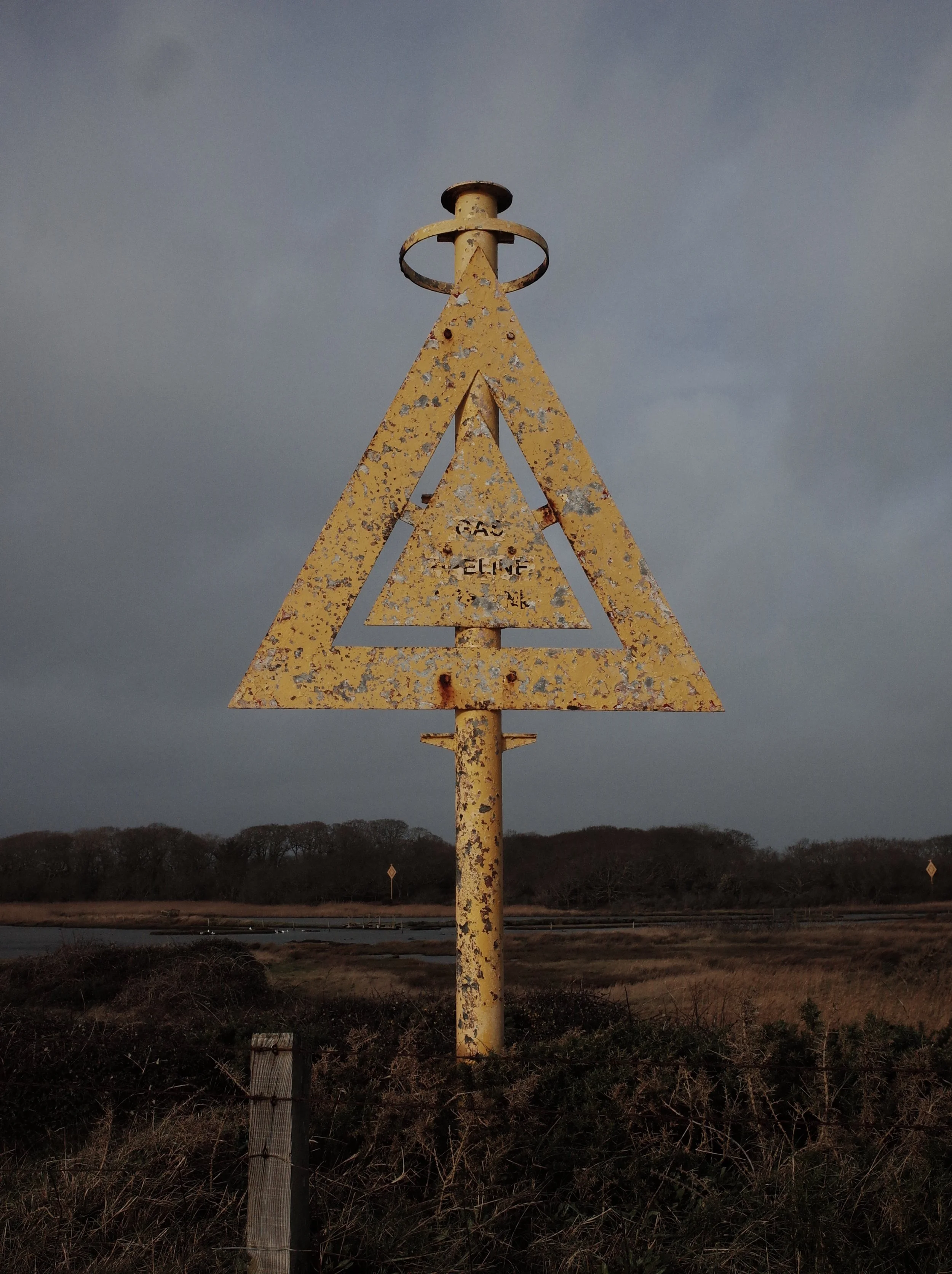 A weathered yellow metal sign in the shape of an upward-pointing triangle with a smaller, hollow triangle inside, mounted on a pole in a rural landscape, with dark cloudy sky overhead.