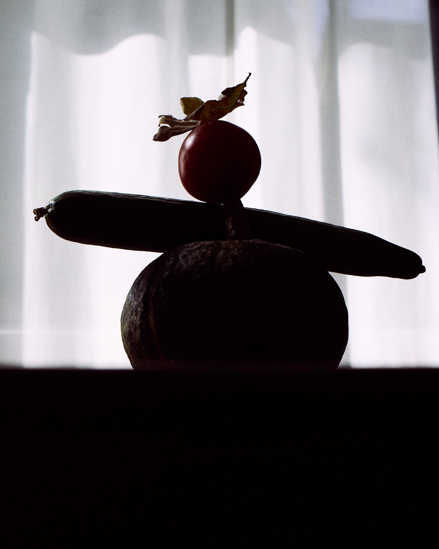 Stacked fruits with tomato, zucchini, lemon, and dried leaf silhouette against sheer curtain background.