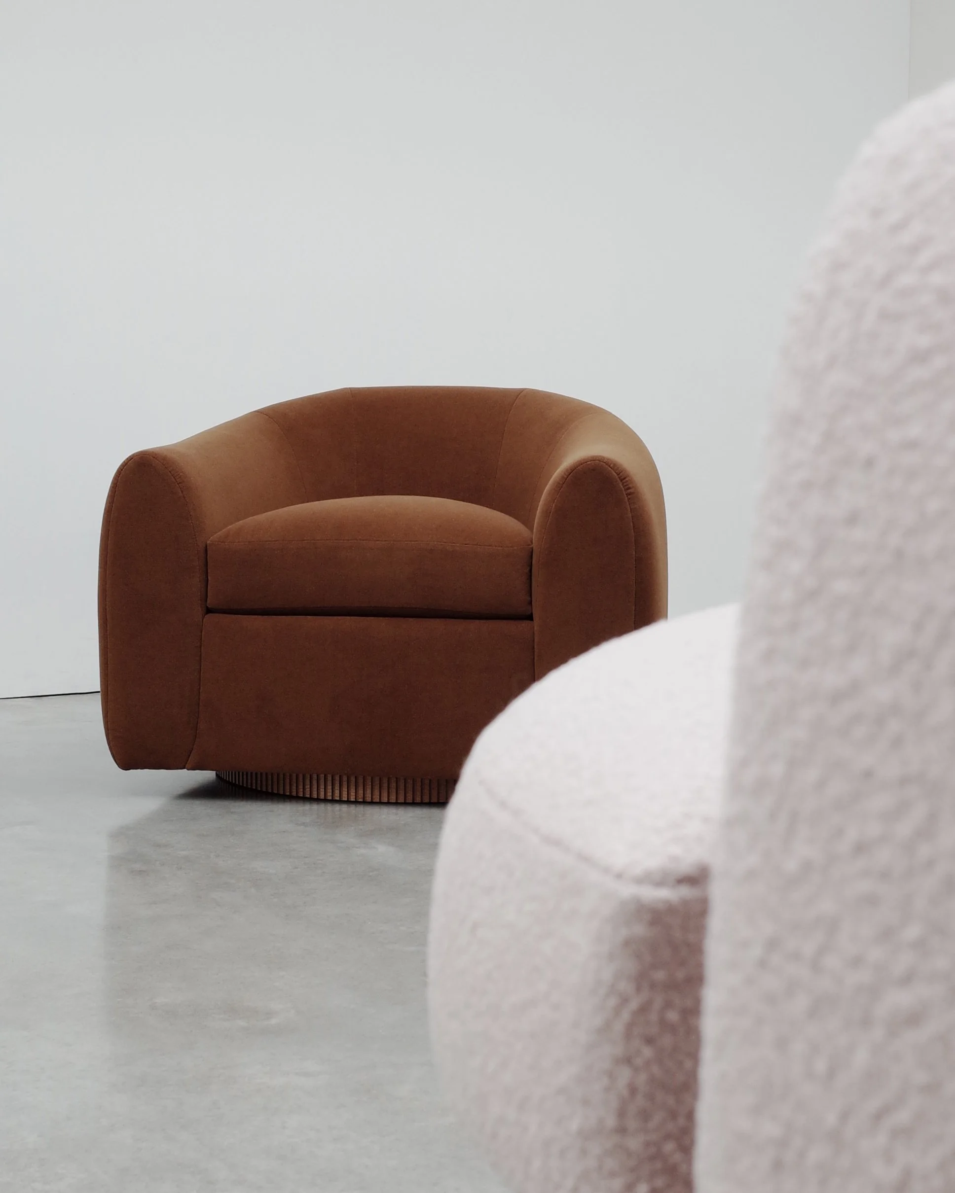 A brown armchair with rounded arms and back, sitting on a grey floor against a plain white wall. Part of a light-colored, textured object is visible in the foreground on the right side.