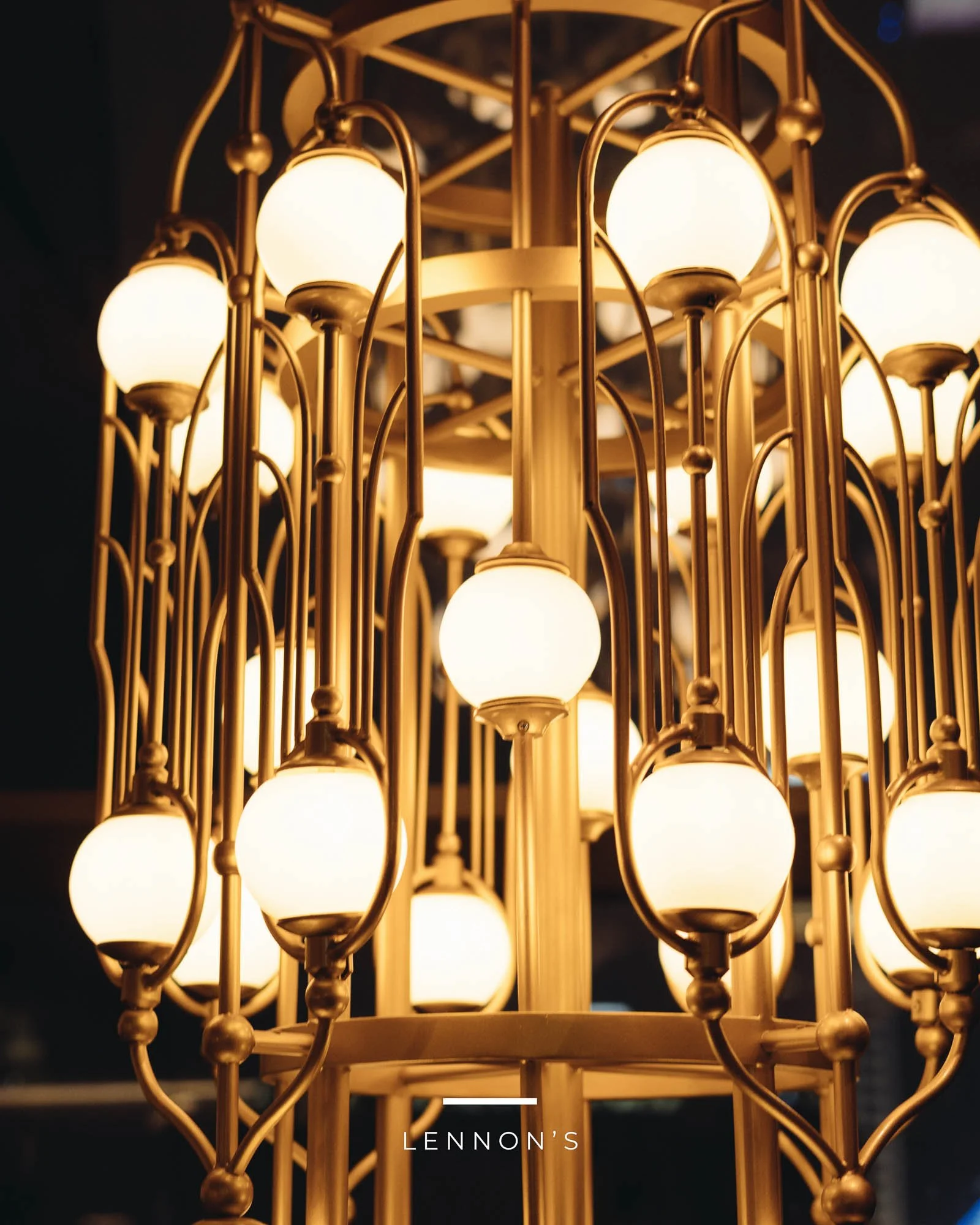 Luxurious gold chandelier with multiple spherical white bulbs hanging from curvilinear gold arms, with the word "LENNON'S" at the bottom.