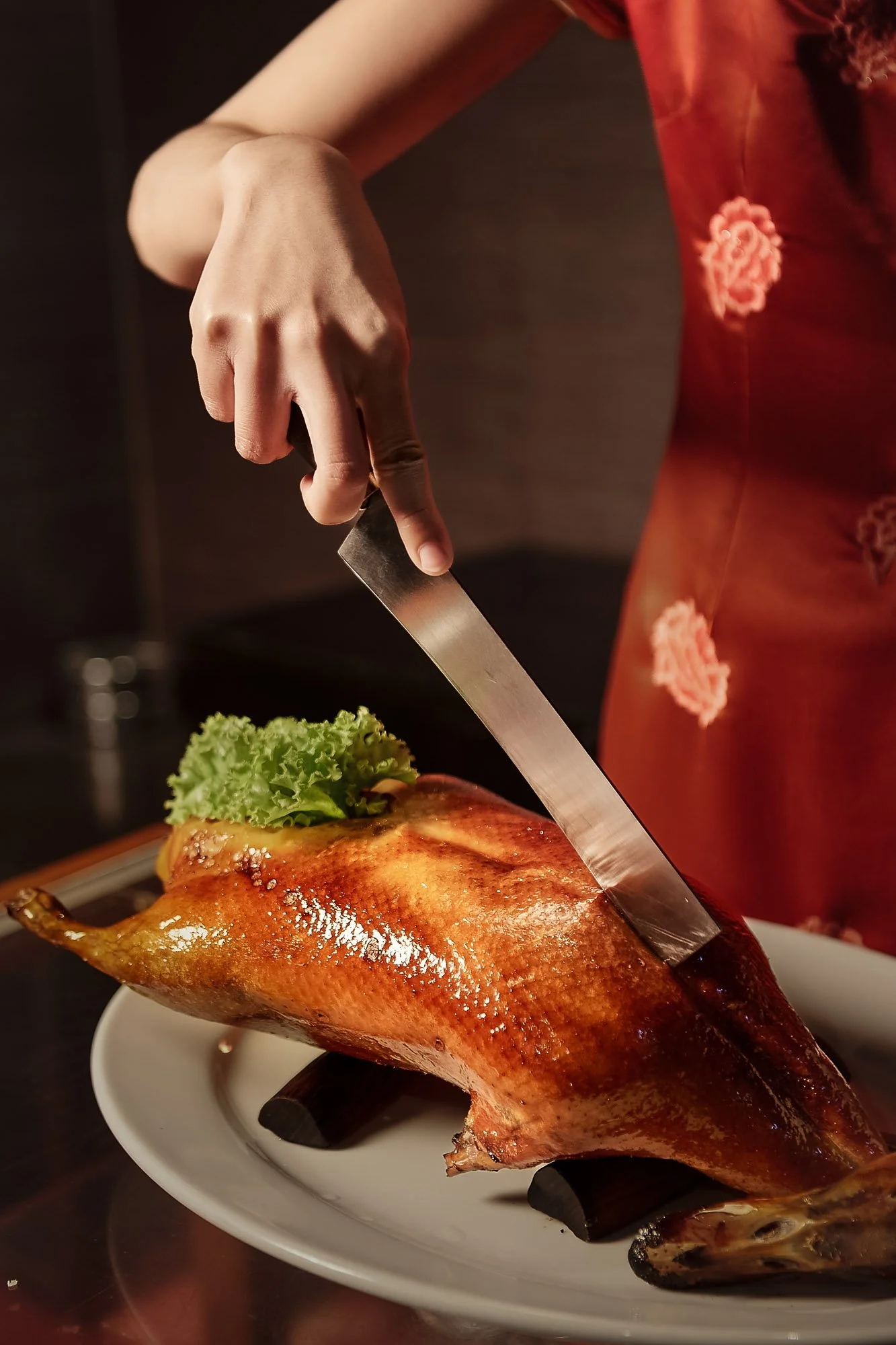 Person in red dress carving a roasted whole duck with a knife on a white plate, garnished with lettuce head.