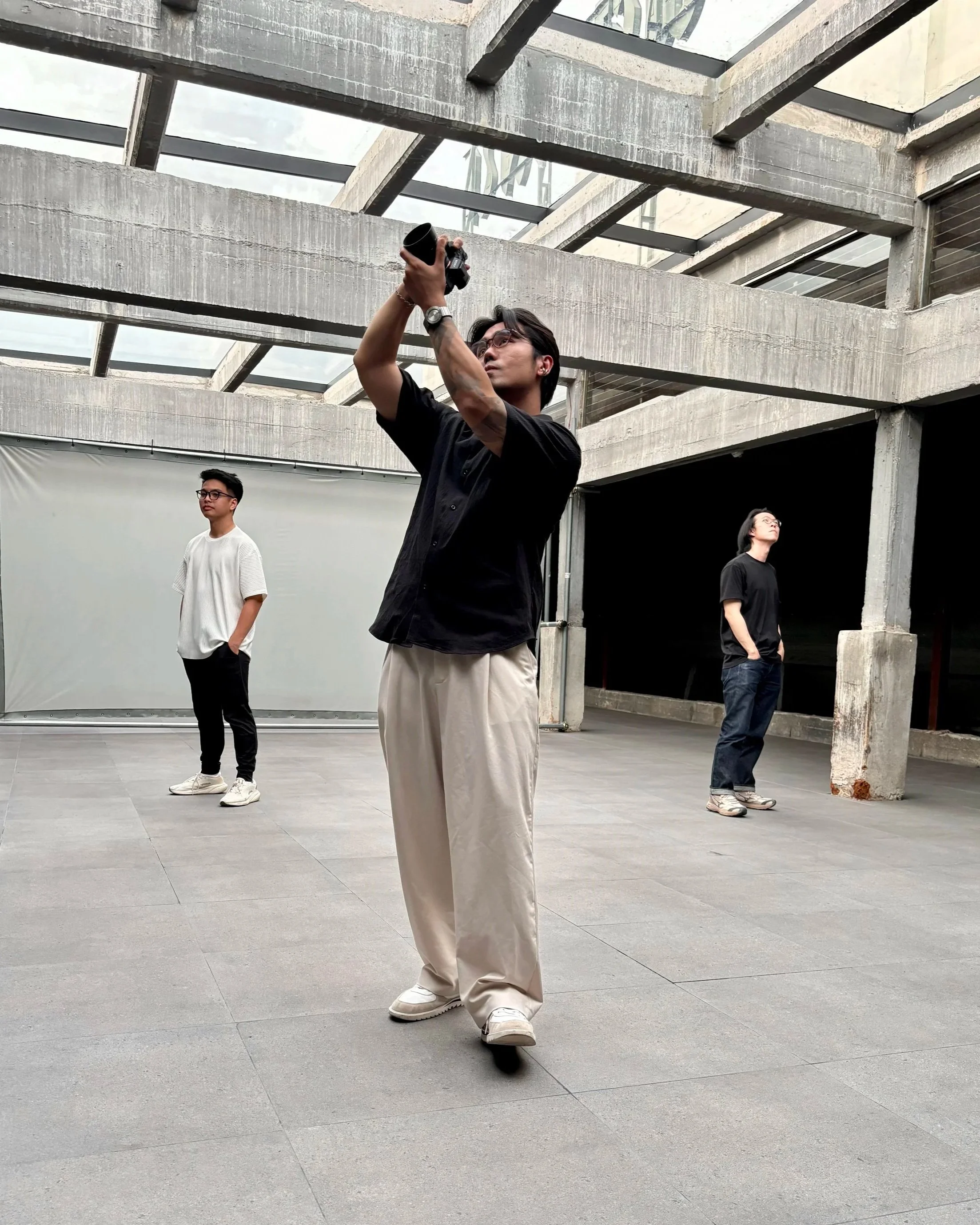 Three young men are standing in an open, modern concrete and glass structure. One man in black is taking photos or videos with a camera, the man in white is standing with his hands in his pockets, and the third man in black is looking upward.