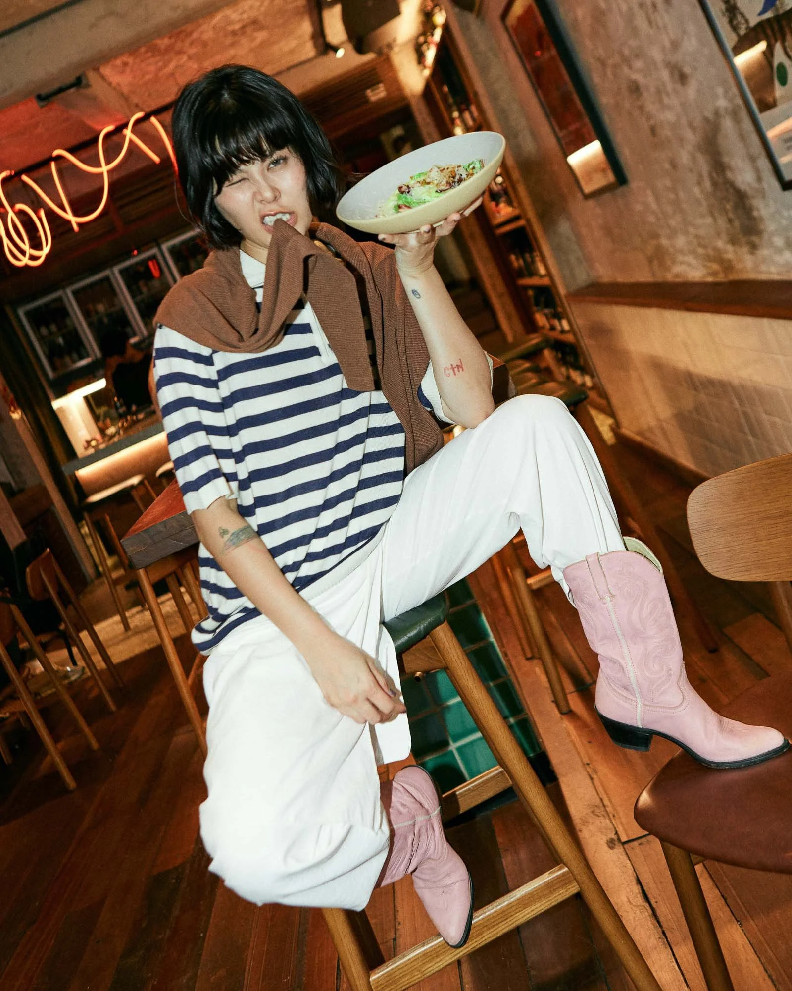 A woman with dark hair, wearing a striped navy and white top, white pants, and pink cowboy boots, sitting on a wooden stool in a restaurant, holding a bowl of salad in one hand, making a playful expression with one eye closed and a slight grin.