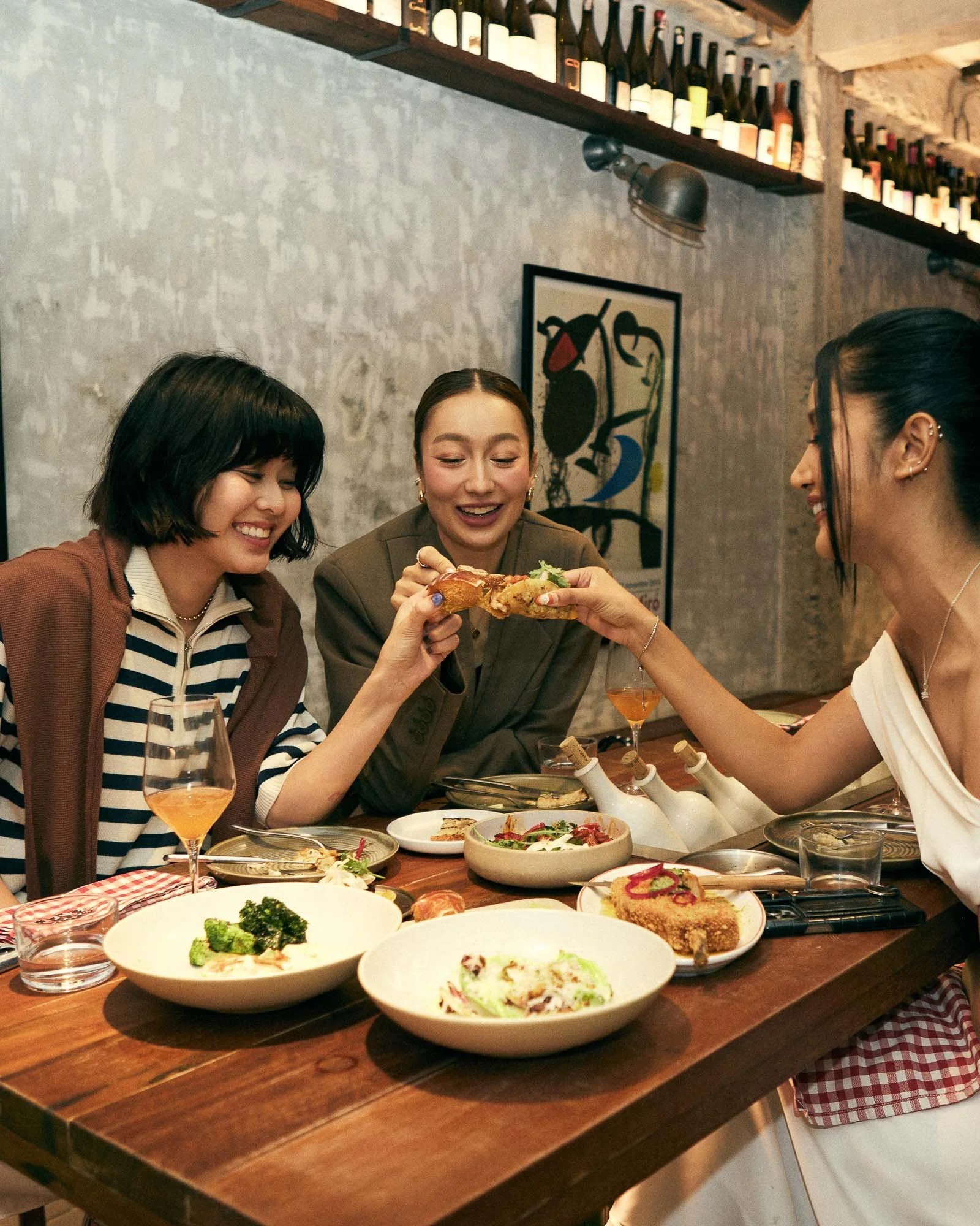Three women sharing a slice of pizza at a restaurant table with various dishes and drinks, smiling and enjoying their meal.