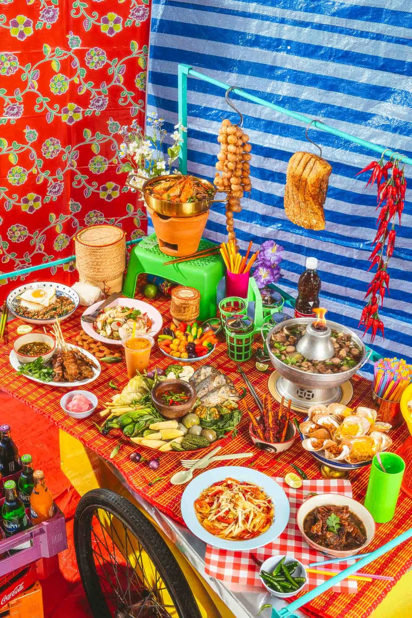 A colorful outdoor food stand with a table full of various Asian dishes, drinks, and condiments, surrounded by vibrant fabric and decorations.
