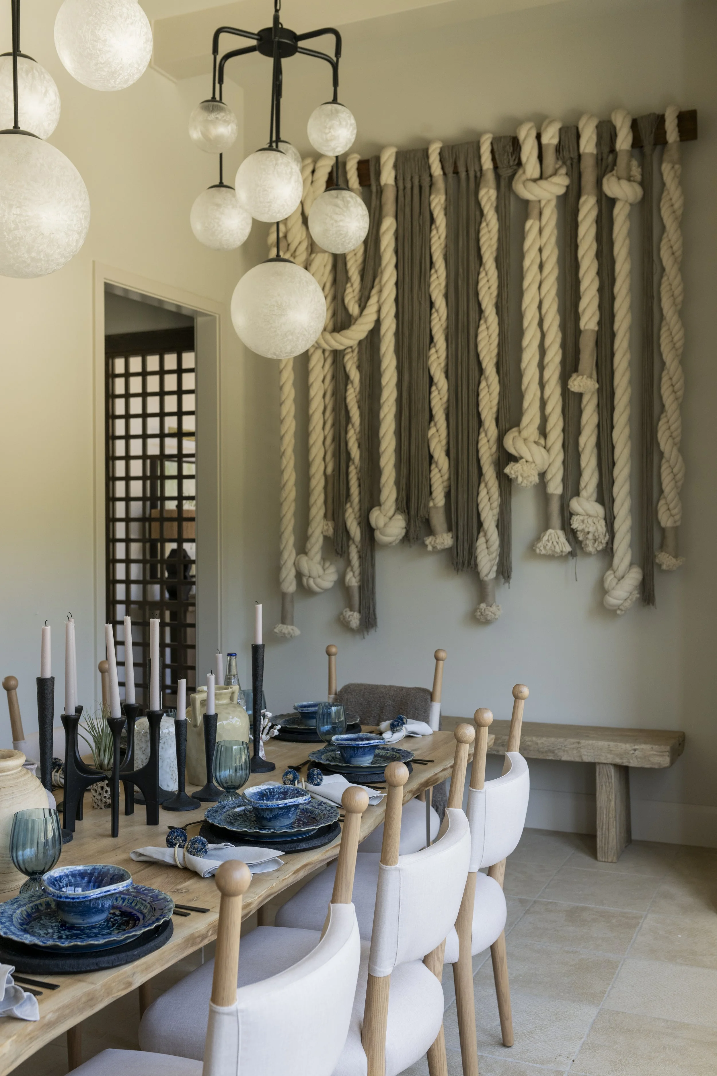 Luxury lake house dining room design with a long wood dining table, blue ceramic place settings, white upholstered chairs, globe pendant lights, and a textured wall hanging.