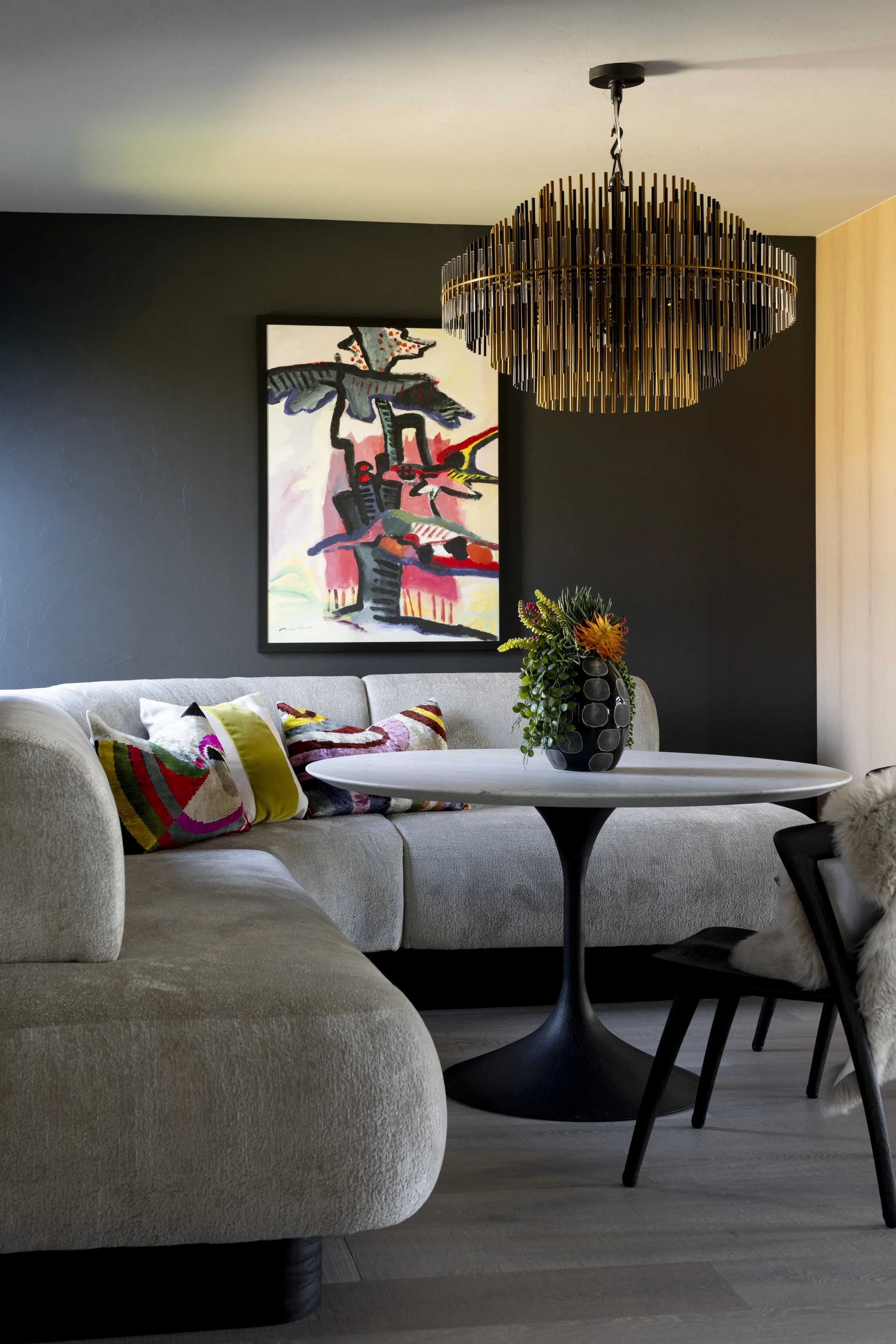 Luxury modern dining nook with a curved cream banquette, round white table, black and gold chandelier, and colorful abstract art against dark walls.