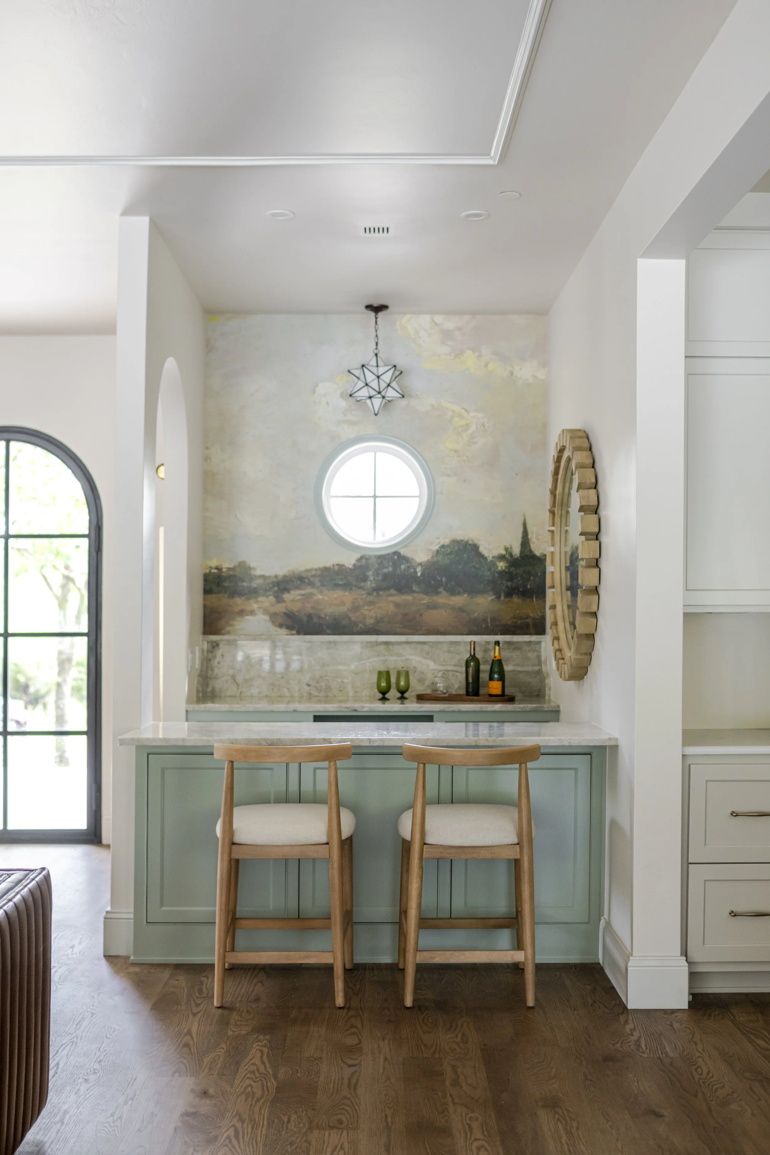 A cozy kitchen nook with mint green cabinetry, two wooden bar stools with cream cushions, a marble countertop, and a landscape mural on the wall featuring trees and a cloudy sky. There are three bottles and two glasses on the counter, a round window above, and modern lighting fixtures.