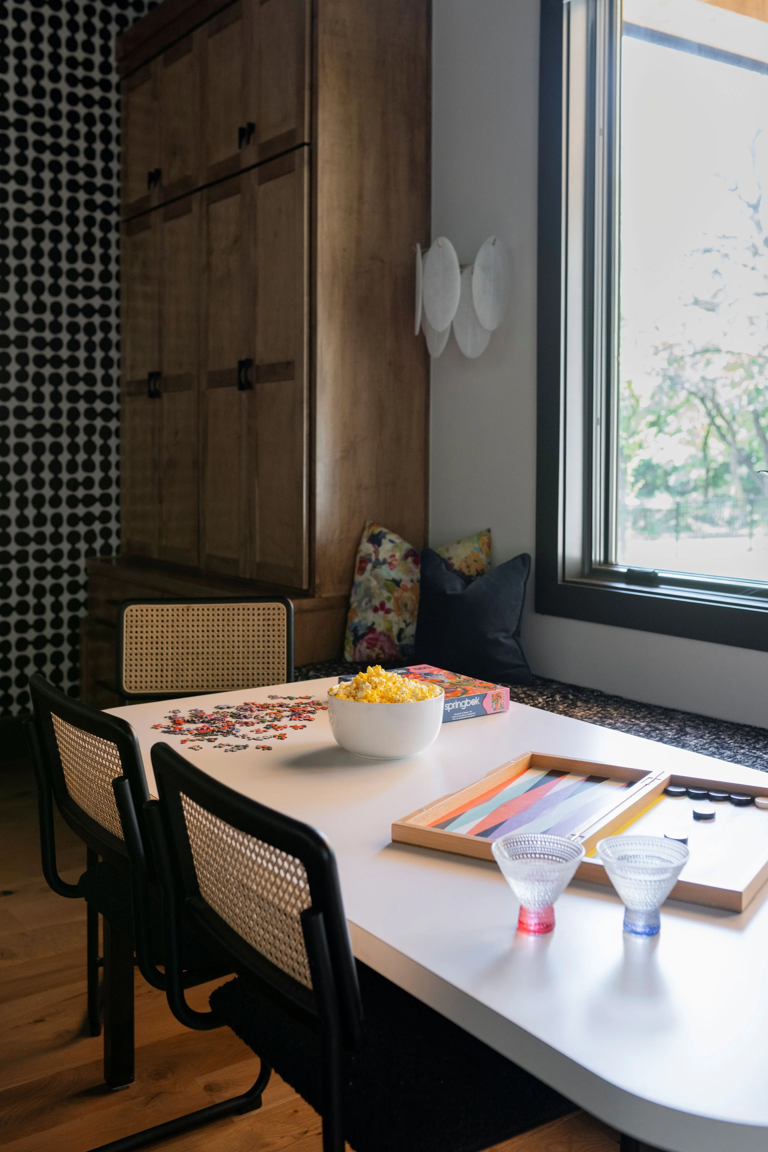 Interior design of a sunlit game room with a white puzzle table, cane chairs, a cushioned window seat, and custom wood cabinetry.