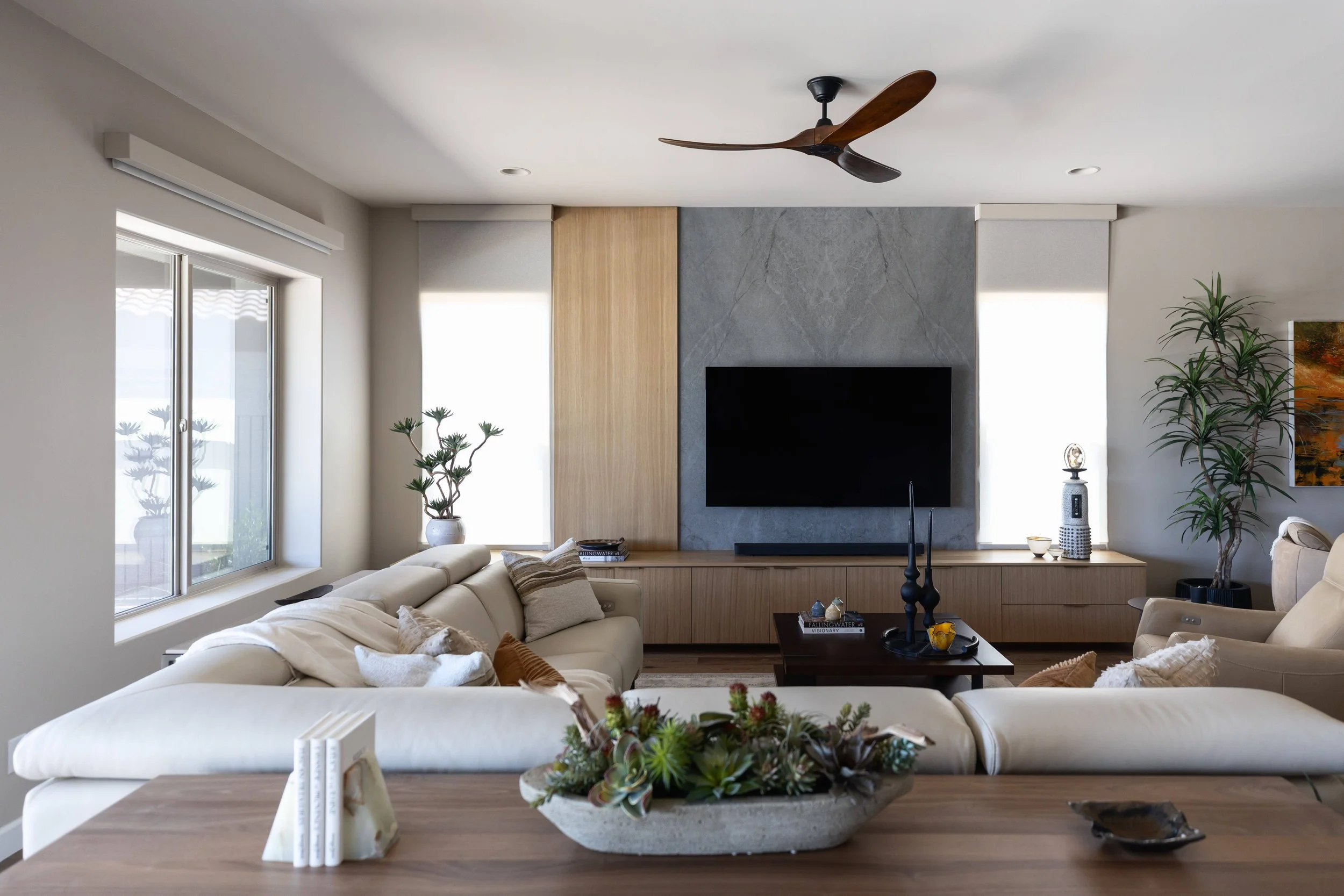 A modern living room with white sofas, plants, a wall-mounted TV, and a ceiling fan.